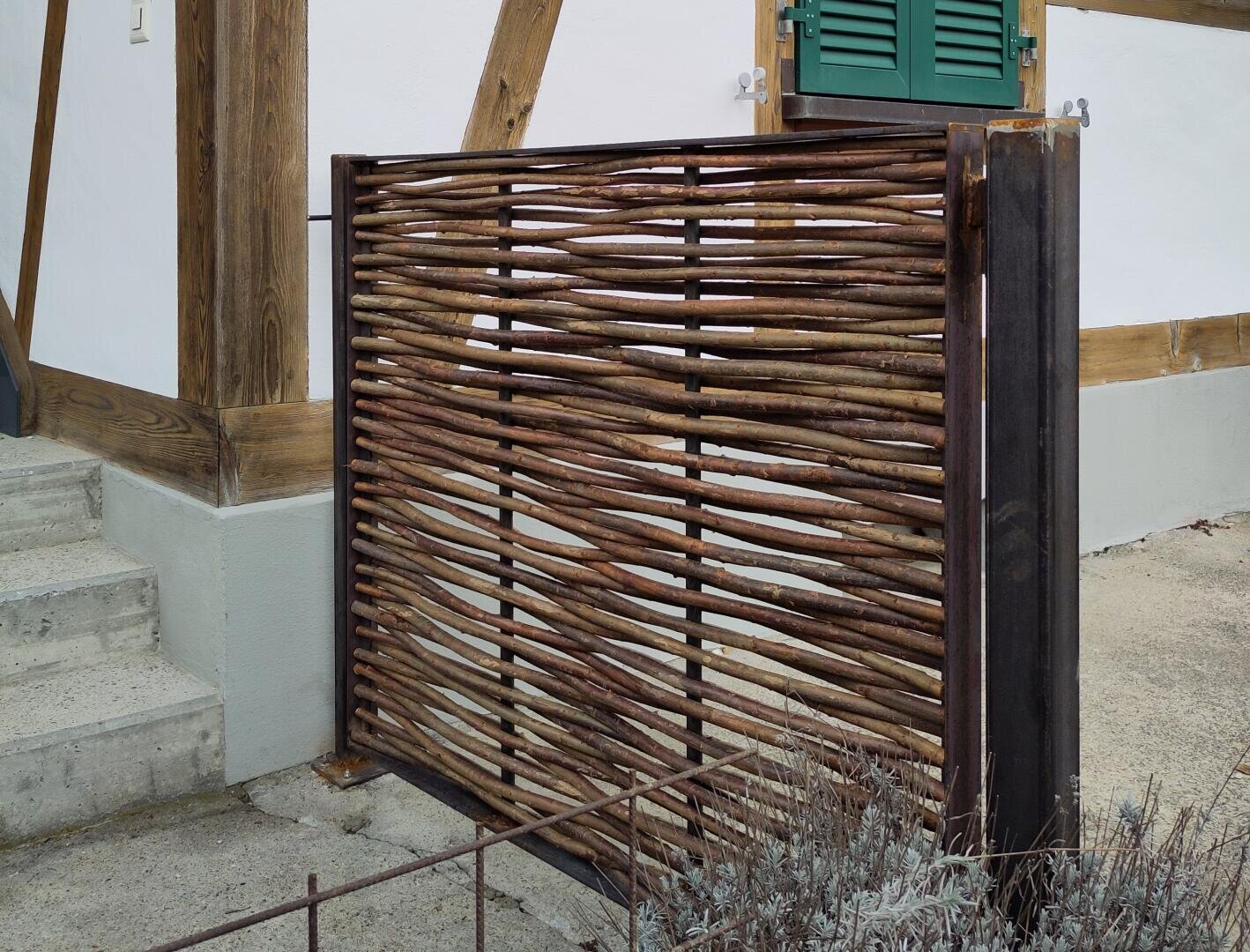 A woven wooden fence with a metal frame stands next to concrete steps in front of a house with white walls, green shutters and exposed wooden beams. Dried plants can be seen in the foreground.