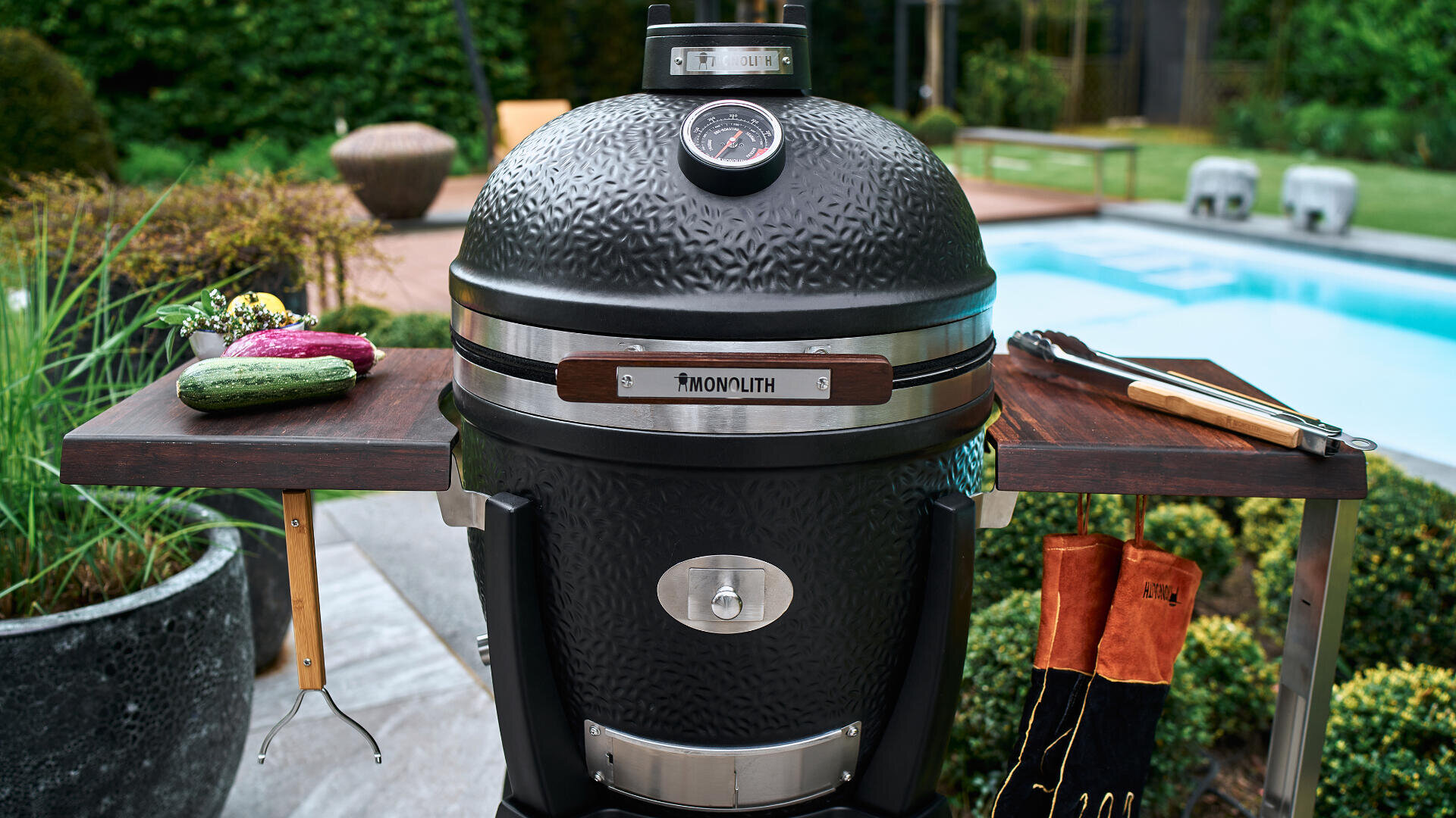Ein schwarzer Keramikgrill mit Temperaturanzeige steht auf einer Terrasse neben einem Pool, mit hölzernen Seitenregalen für frisches Gemüse, Grillwerkzeug und hitzebeständige Handschuhe. Im Hintergrund sind Grünpflanzen und Gartenmöbel zu sehen.