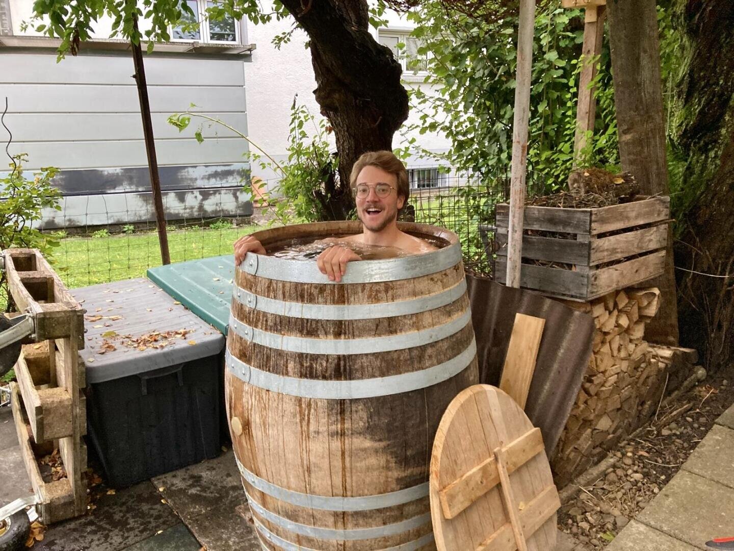 A smiling man with glasses sits in a large wooden barrel filled with water in a backyard, surrounded by trees, firewood, wooden crates and a gray building in the background.