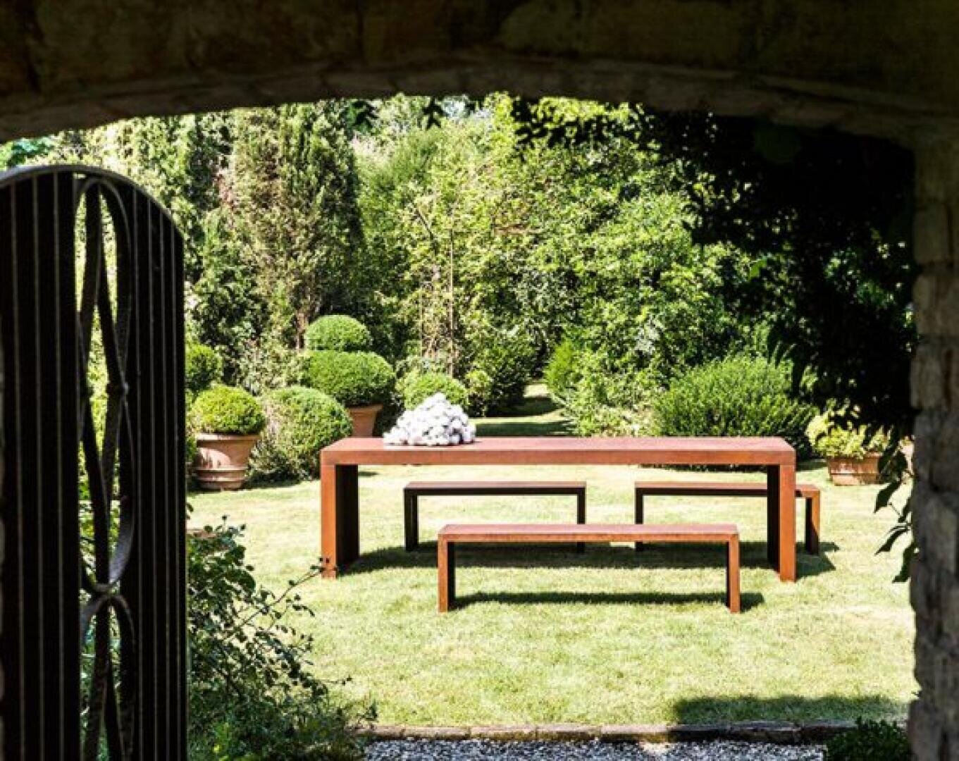 A modern wooden table with benches sits on a grassy lawn, surrounded by potted plants and lush greenery, viewed through an open gate and stone archway.