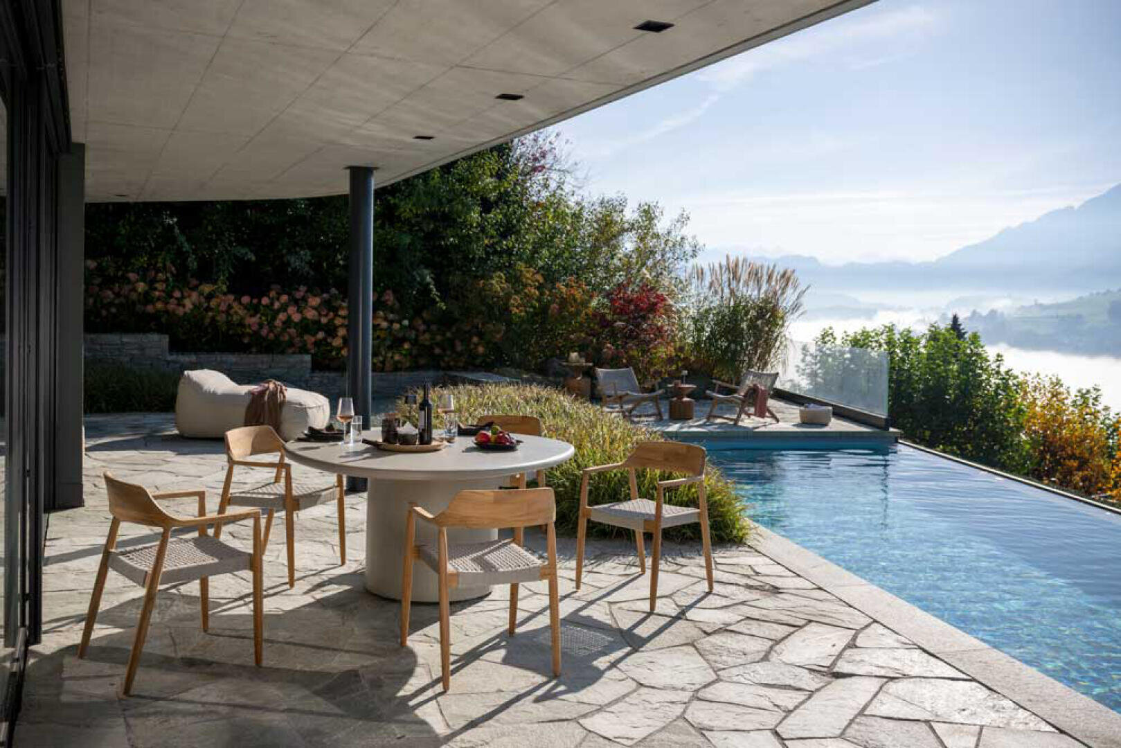 Eine moderne Terrasse mit rundem Tisch und Holzstühlen blickt auf einen Infinity-Pool. Zwei Personen entspannen am Pool inmitten von üppigem Grün, mit Bergen und einem nebligen Tal im Hintergrund unter einem klaren Himmel.