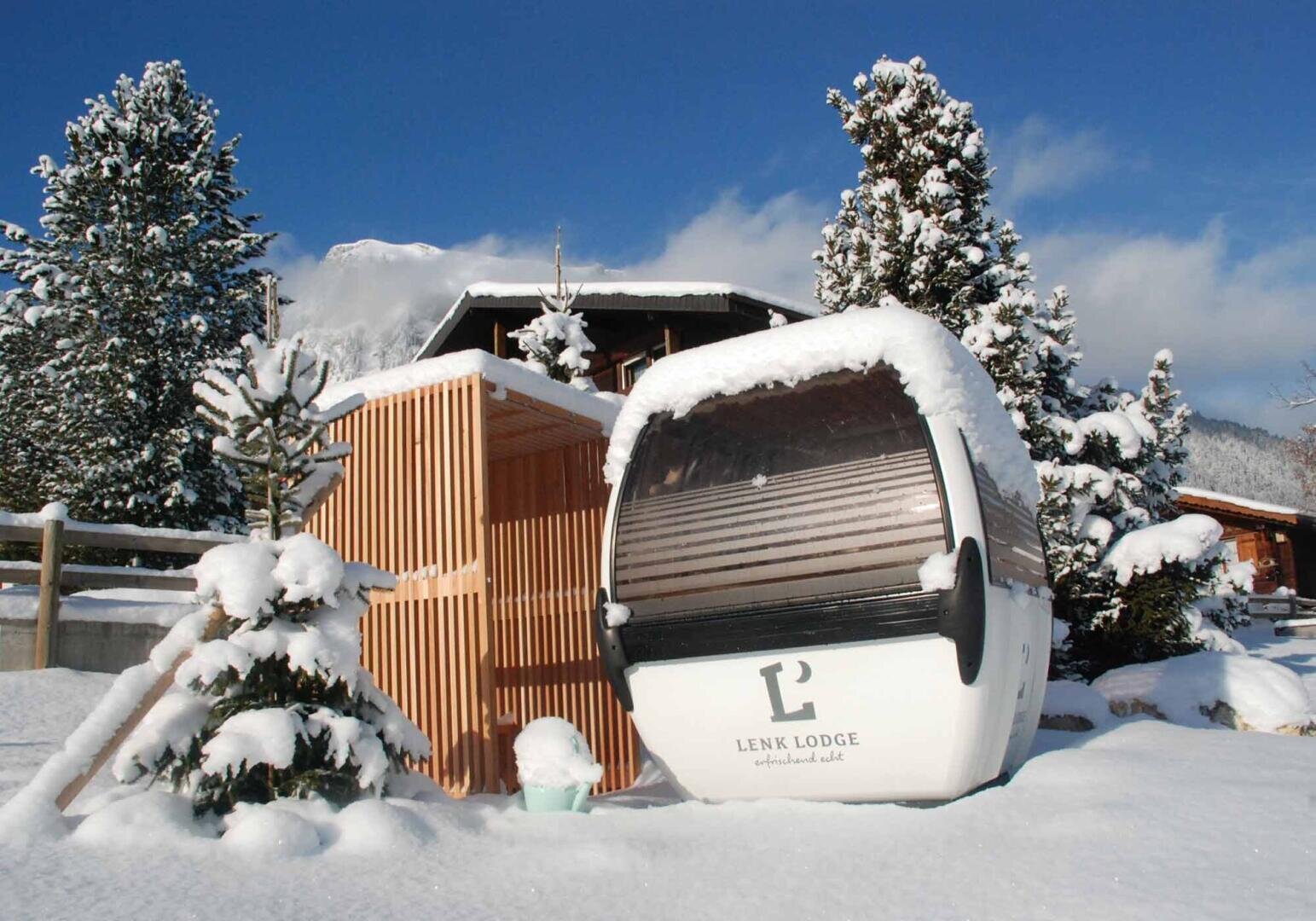Eine verschneite Szene mit einer kleinen Gondelhütte mit der Aufschrift Lenk Lodge neben einer Holzkonstruktion, umgeben von schneebedeckten Bäumen und Bergen im Hintergrund.
