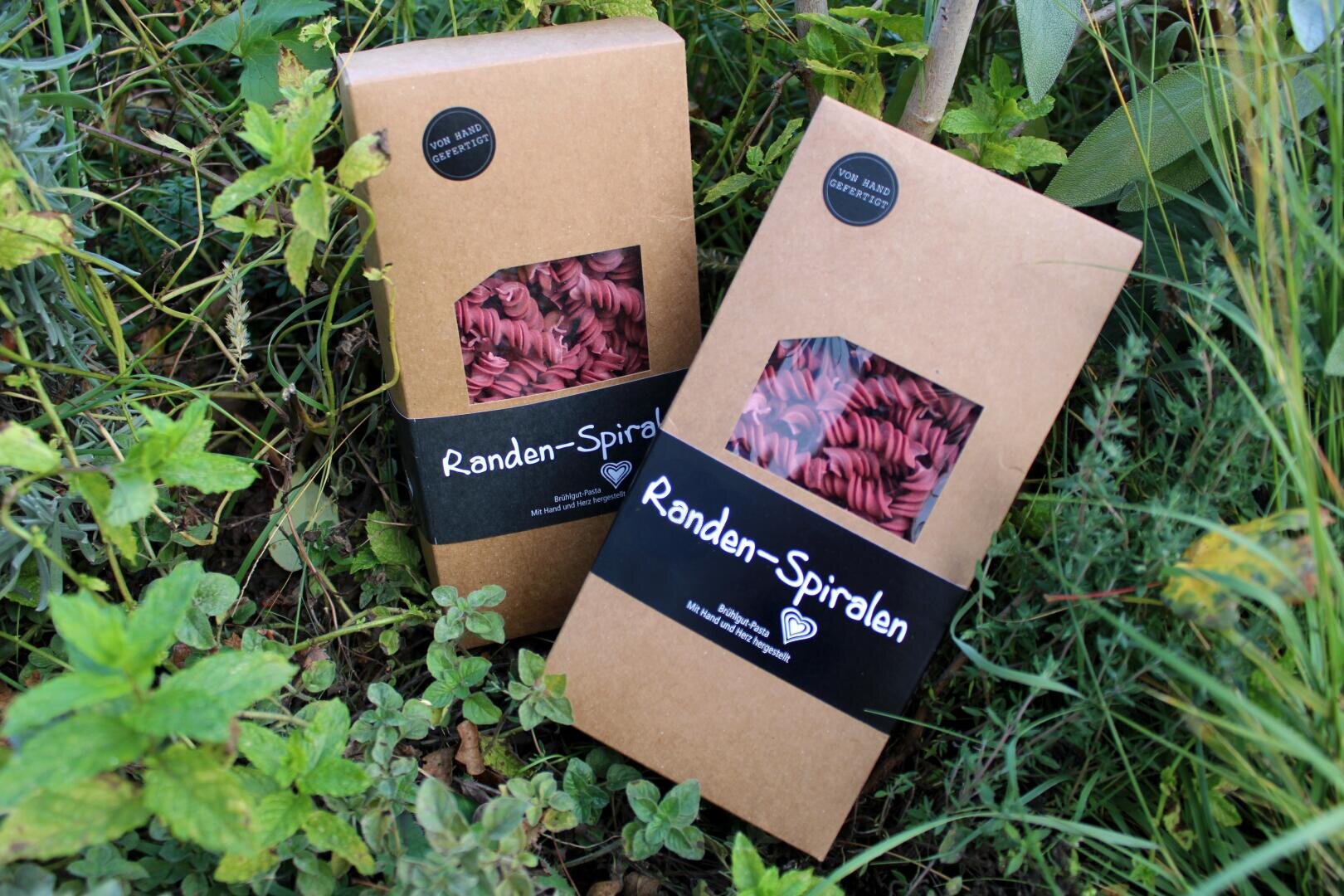 Two cardboard boxes of "beetroot spirals" with a window in which pink noodles can be seen are standing outside among the green plants and grass.