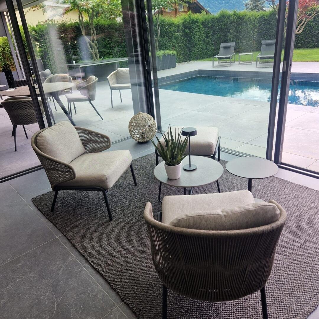 Moderner Sitzbereich im Innenbereich mit zwei Sesseln, zwei kleinen runden Tischen und einer Pflanze auf einem Teppich vor großen Glasfenstern mit Blick auf eine Terrasse und einen Pool mit Liegestühlen und Grünpflanzen im Freien.