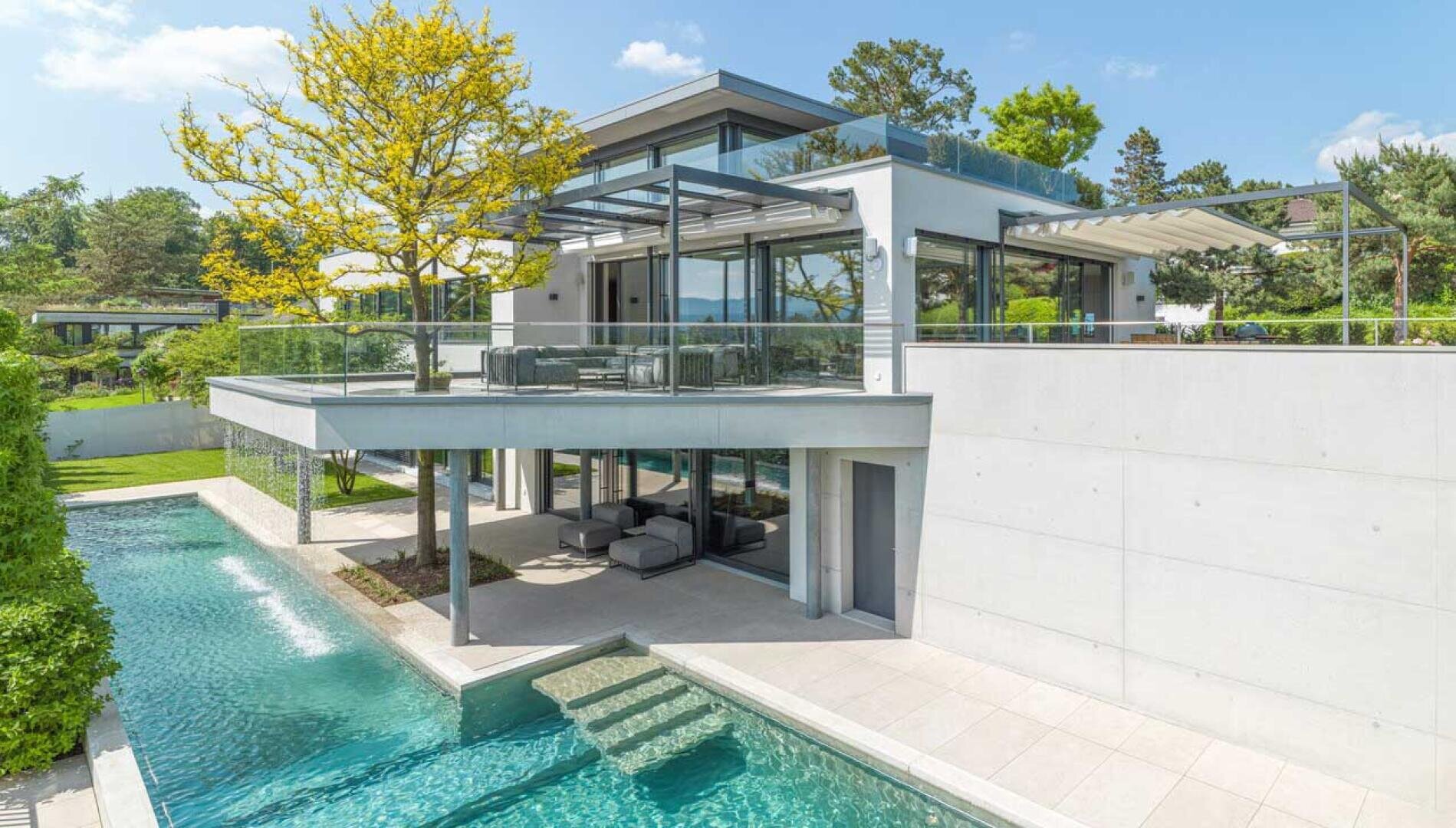 Ein modernes zweistöckiges Haus mit großen Glasfenstern, geräumigen Balkonen und einem Swimmingpool, umgeben von Grün unter einem klaren blauen Himmel.