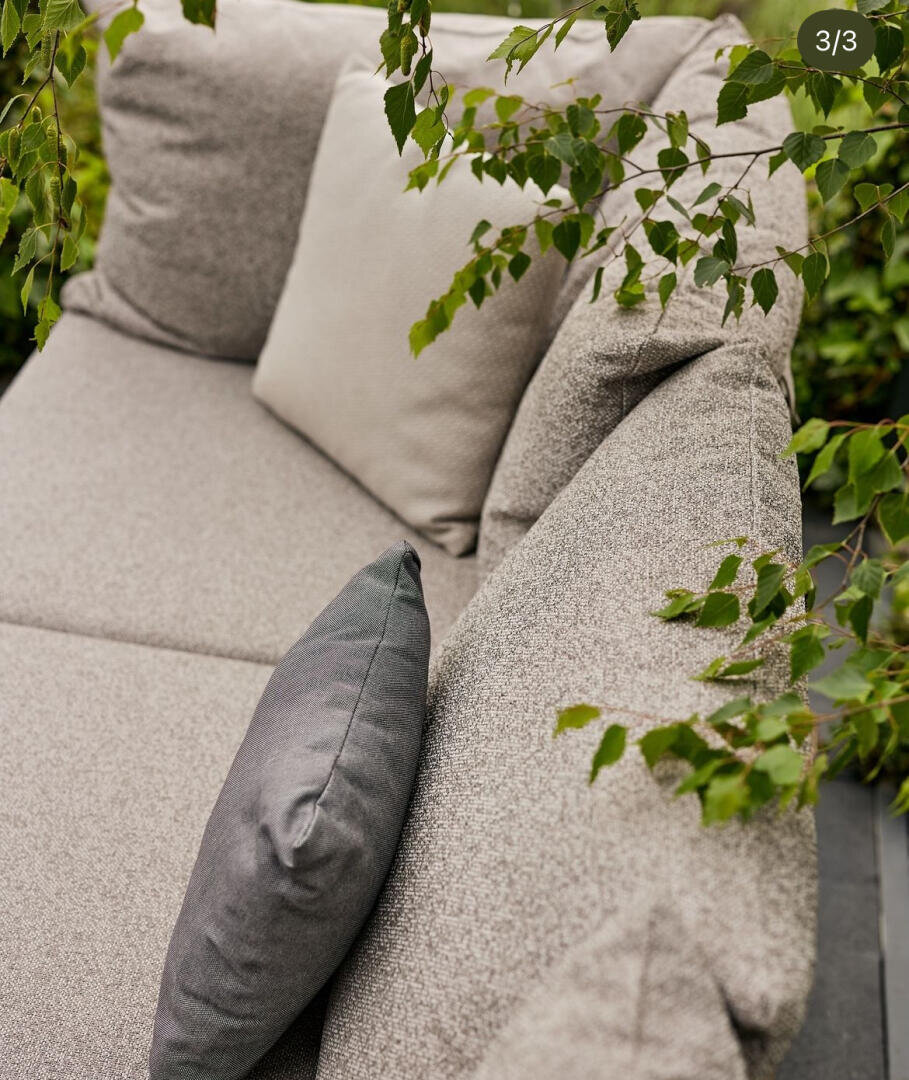 Nahaufnahme eines hellgrauen Sofas im Freien mit zwei Kissen, einem hell- und einem dunkelgrauen, die teilweise von überhängenden grünen Blättern bedeckt sind.