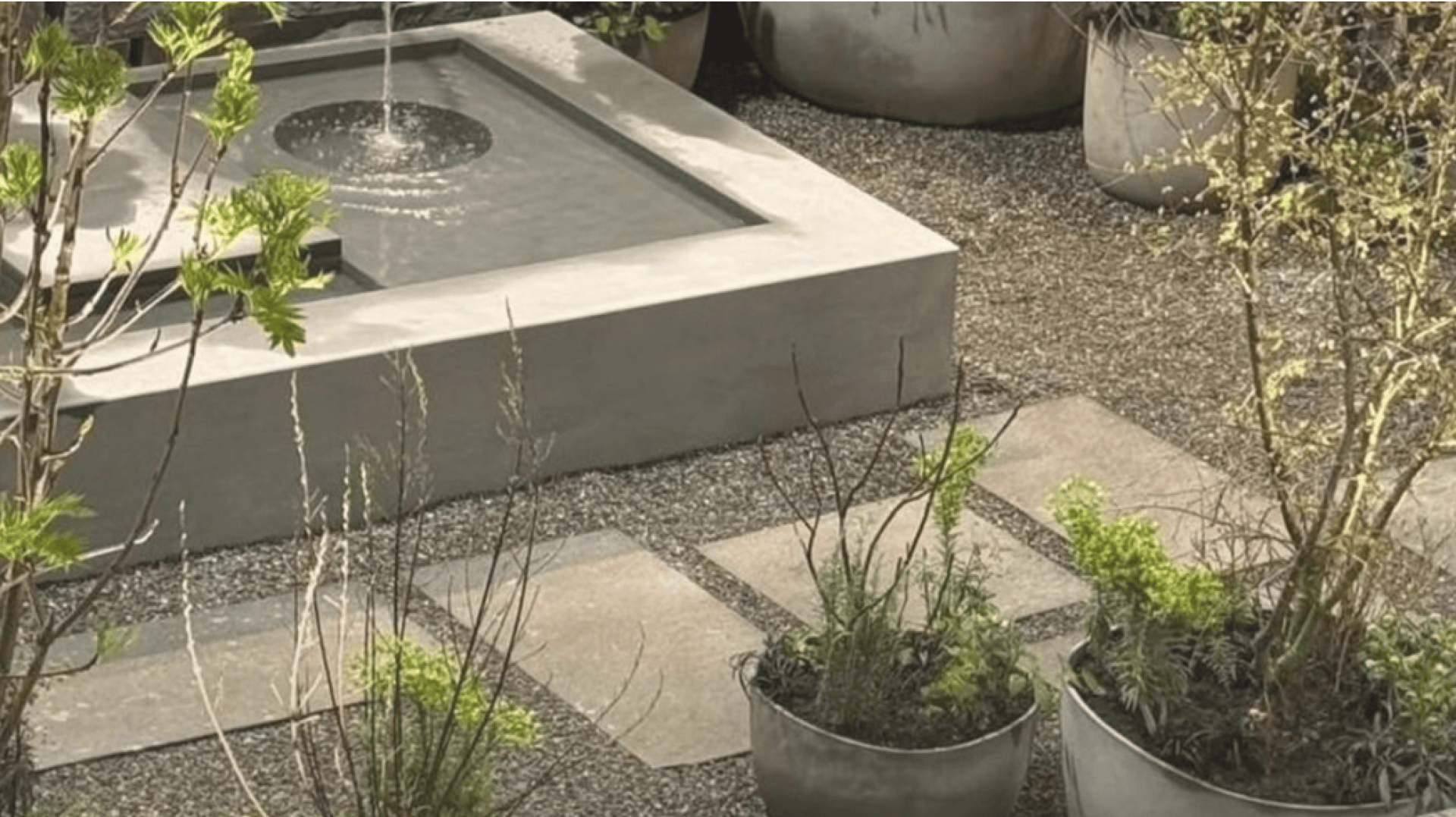 A modern square water fountain with a central spout is surrounded by gravel, stone pavers, and potted green plants in a garden setting.