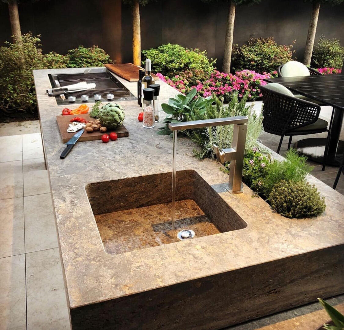 Eine moderne Außenküche verfügt über eine Arbeitsplatte aus Stein mit einem Waschbecken, einem laufenden Wasserhahn, frischem Gemüse, Kräutern und Schneidebrettern. Um die Küche herum sind Grünpflanzen, Blumen und ein Essbereich mit dunklen Stühlen.