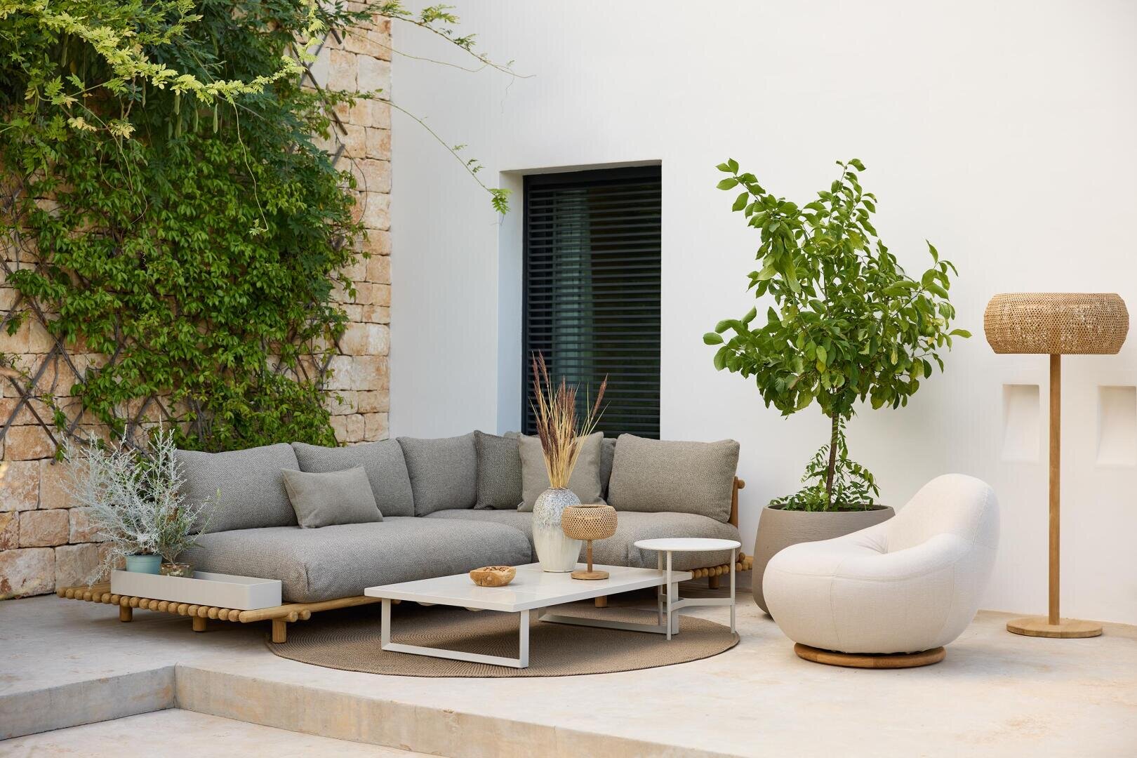 Eine stilvolle Außenterrasse mit einem grauen Modulsofa, einem weißen runden Stuhl, einem weißen Couchtisch mit Dekoration, Topfpflanzen, einer geflochtenen Stehlampe und steinernen und weißen Wänden mit grünen Ranken.