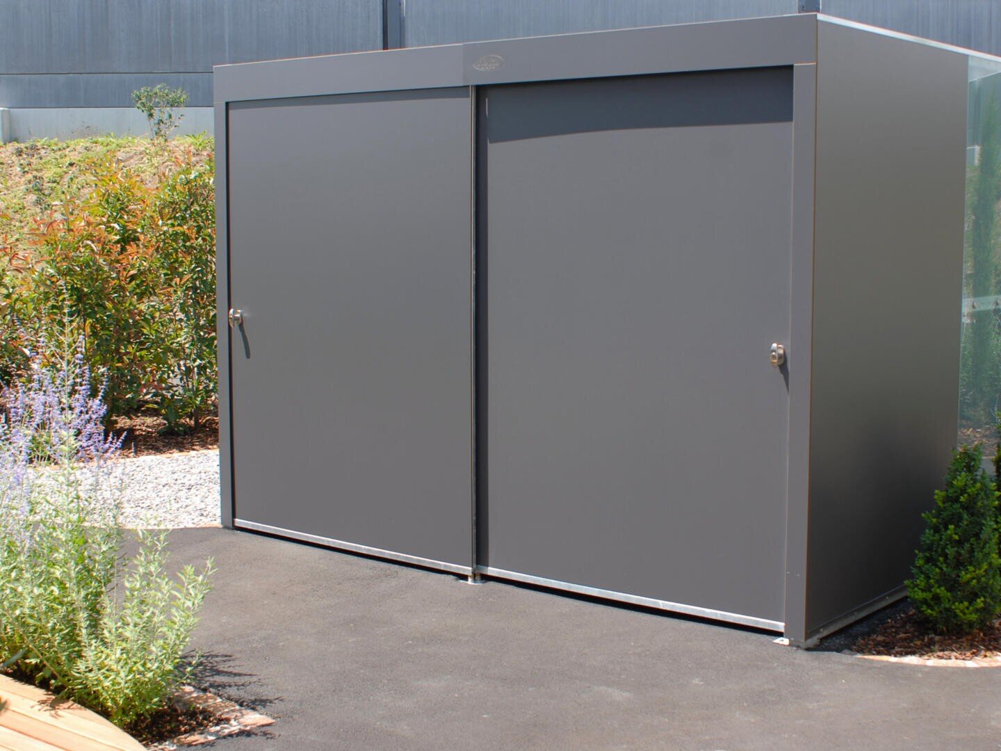 Ein großer, grauer Metallschuppen mit zwei verschlossenen Türen steht auf einer gepflasterten Fläche, umgeben von Grünpflanzen und Kieselsteinen.