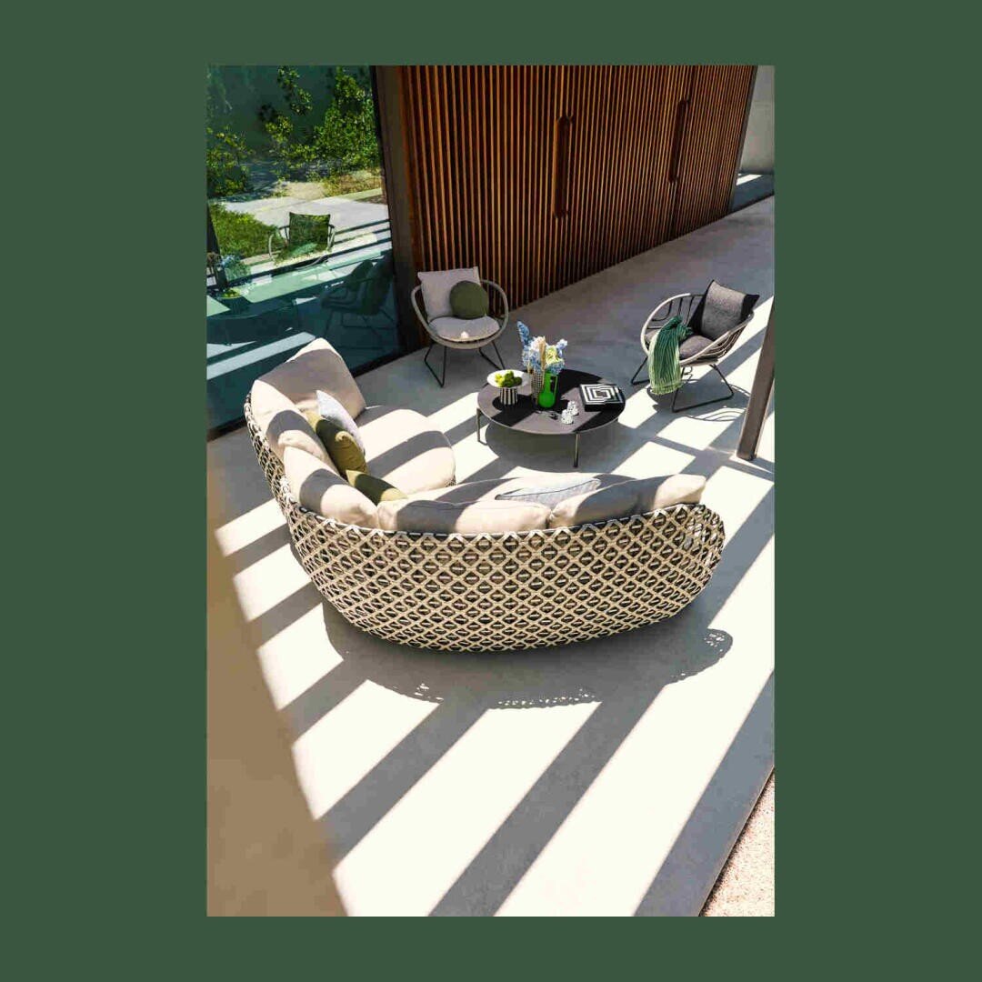 Terrasse im Freien mit einem geschwungenen gemusterten Sofa, zwei Stühlen und zwei runden Tischen auf einer sonnenbeschienenen Terrasse. Im Hintergrund sind Holzlatten und Grünpflanzen zu sehen, wobei die Schatten auf dem Boden gestreifte Muster erzeugen.