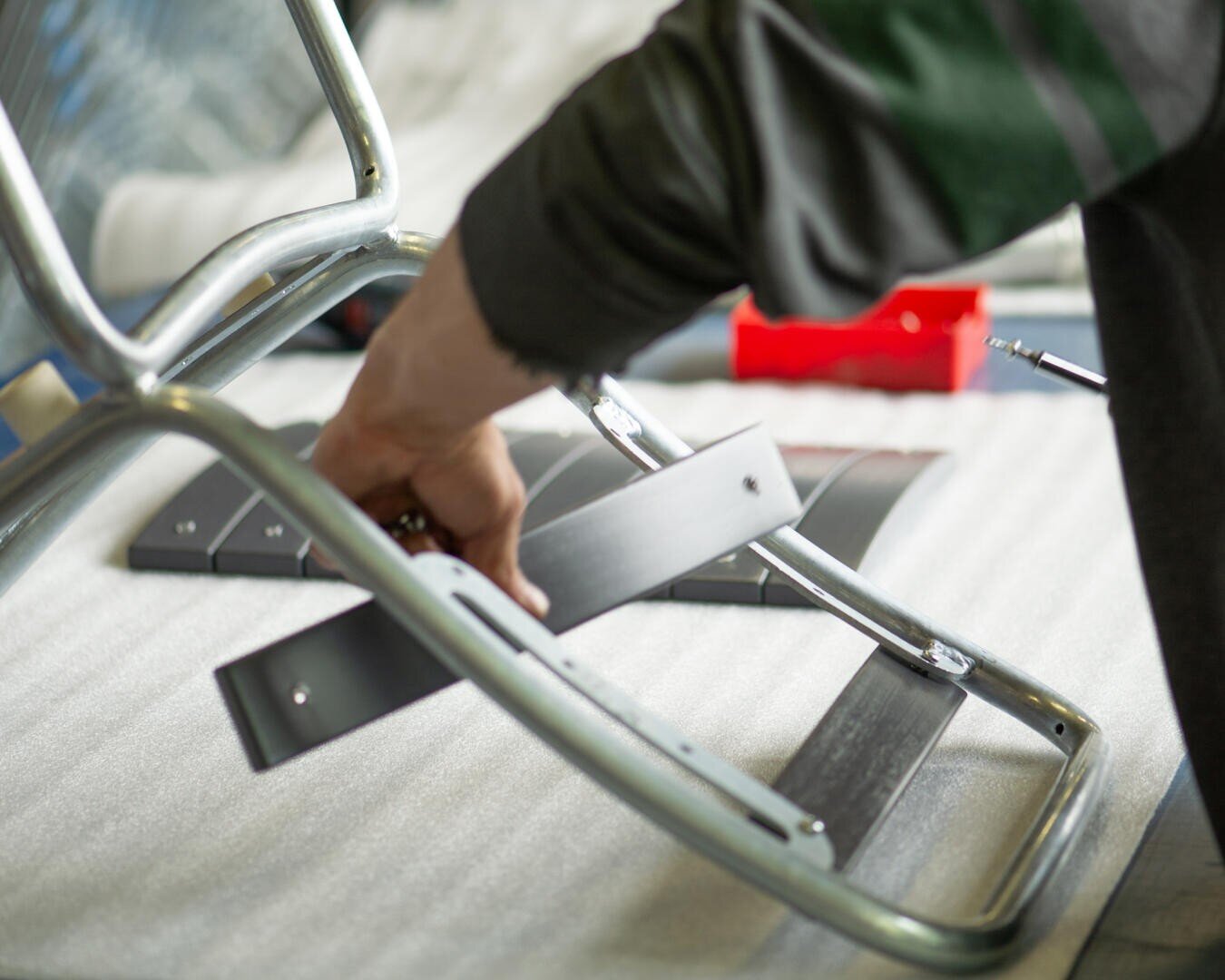 Die Hand einer Person setzt einen Stuhlrahmen aus Metall und Holz zusammen, wobei sie eine schwarze Latte gegen ein silbernes Metallrohr auf einer Arbeitsfläche hält, auf der sich Werkzeuge und Eisenwaren befinden.