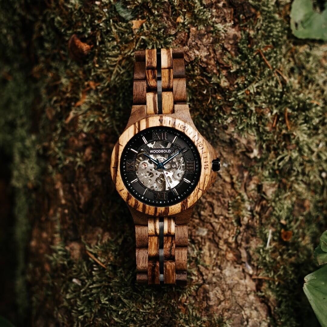 Eine hölzerne Armbanduhr mit sichtbarem mechanischem Uhrwerk ist auf moosbewachsener Baumrinde platziert, wobei sich natürliche Holztöne mit komplizierten Zahnrädern und schwarzen Akzenten verbinden.