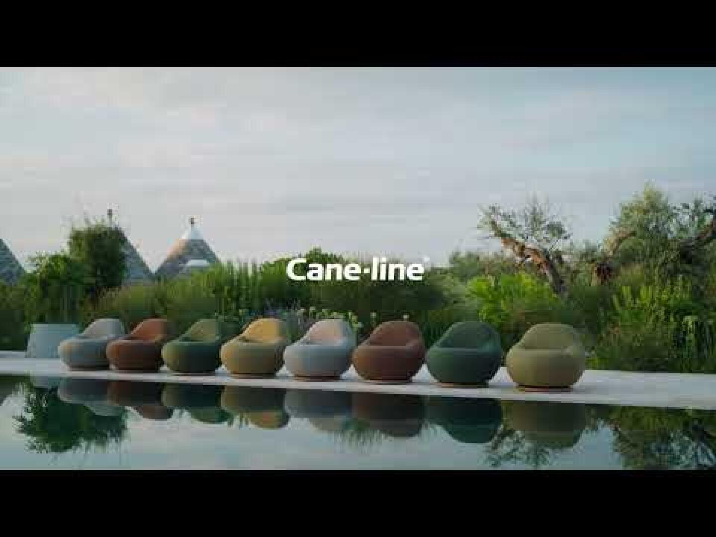 A row of modern, colorful outdoor lounge chairs sits beside a reflective pool surrounded by lush greenery, with trees and pointed-roof buildings in the background. The word Cane-line appears at the center of the image.
