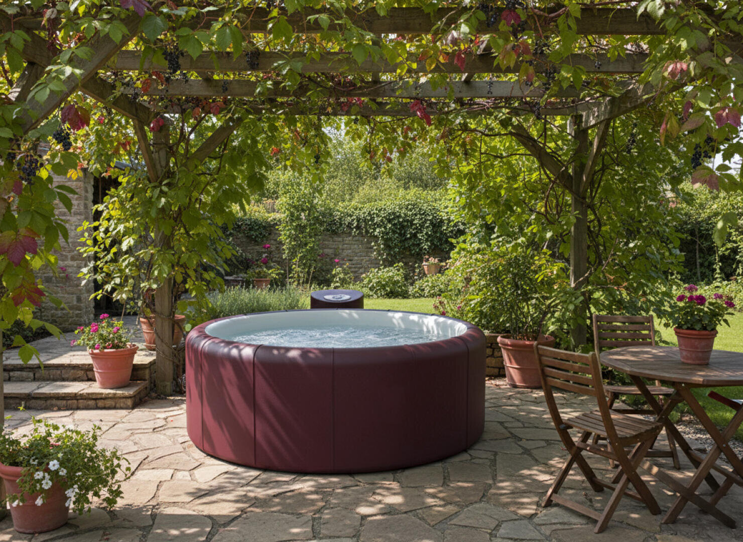 A round, wine-red hot tub stands on a stone terrace under a leafy pergola, surrounded by potted plants. Nearby, a wooden table with chairs stands in the shade, while a lush garden can be seen in the background.