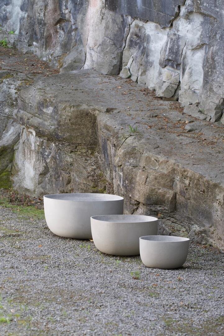 Drei runde, weiße, minimalistische Pflanzgefäße unterschiedlicher Größe stehen in einer Reihe auf einer Kiesfläche neben einer zerklüfteten, felsigen Steinmauer.