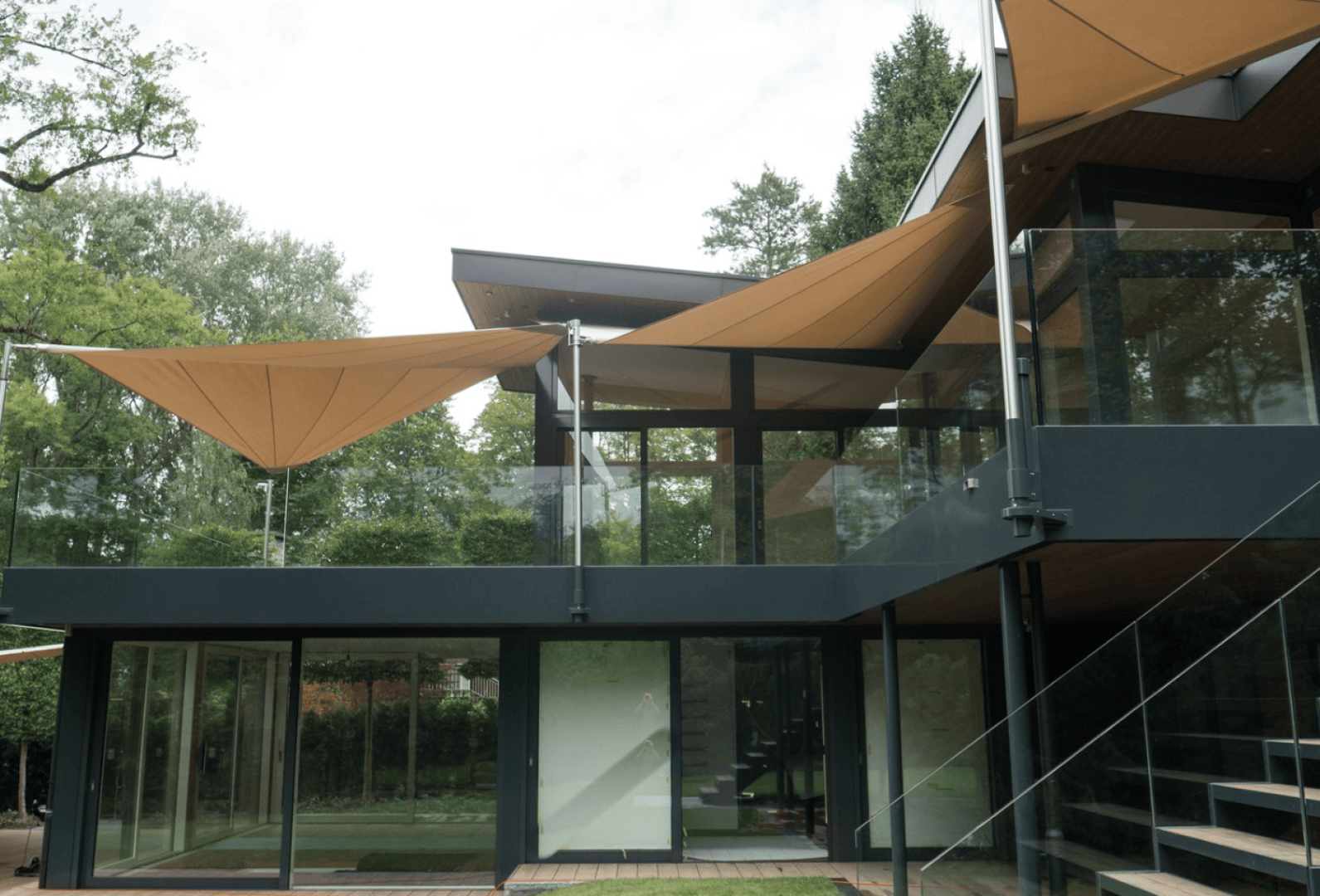 Modernes zweistöckiges Haus mit großen Glasfenstern und -türen, mit beigen dreieckigen Sonnensegeln über der Terrasse. Umgeben von Grün und Bäumen, mit Metallgeländern und einer sichtbaren Außentreppe.