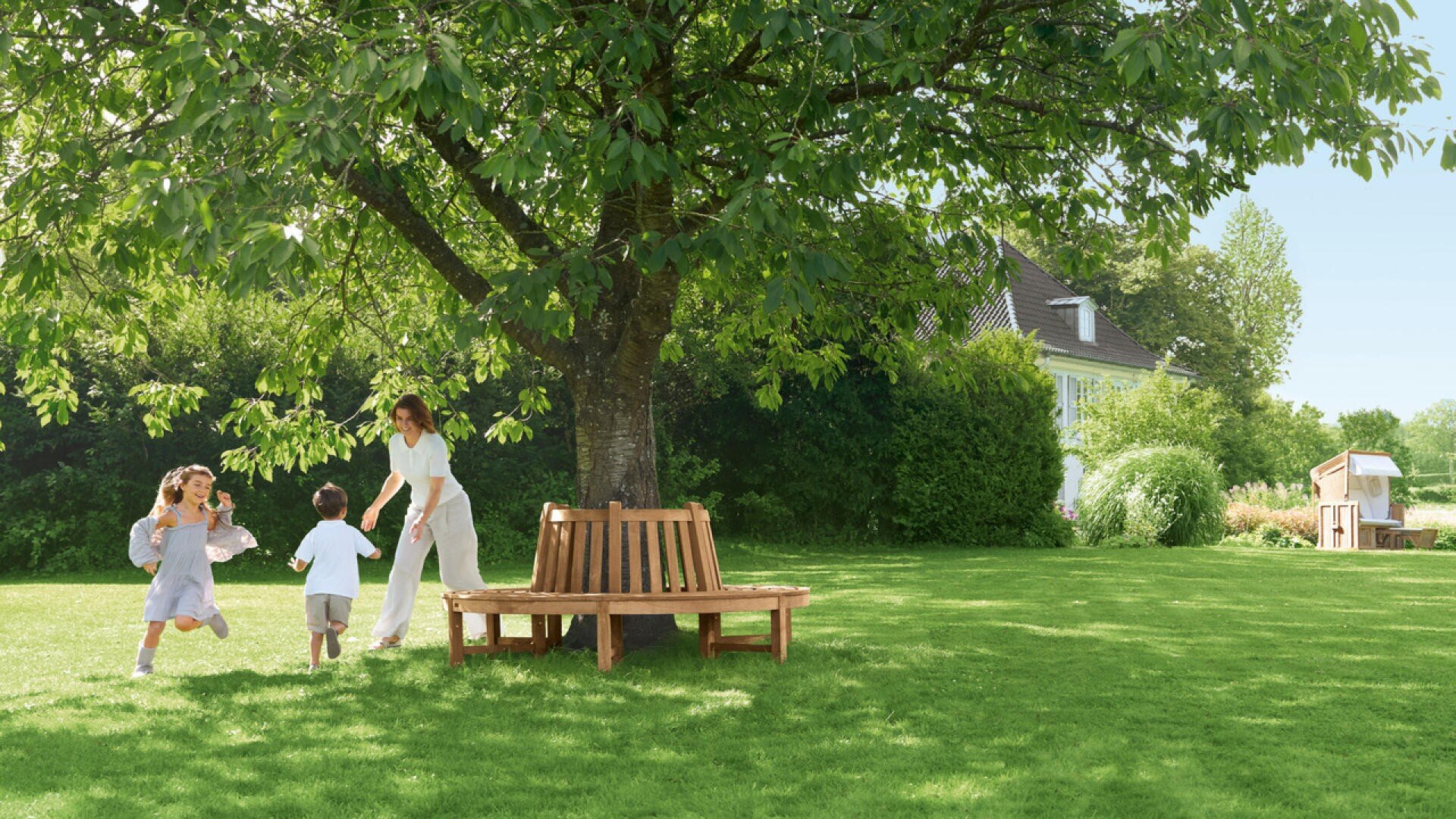 Eine Frau und zwei Kinder spielen auf einem sonnigen, grünen Rasen in der Nähe eines großen Baumes mit einer runden Holzbank. Im Hintergrund sind bei klarem Himmel ein Haus und ein Gartenhaus zu sehen.