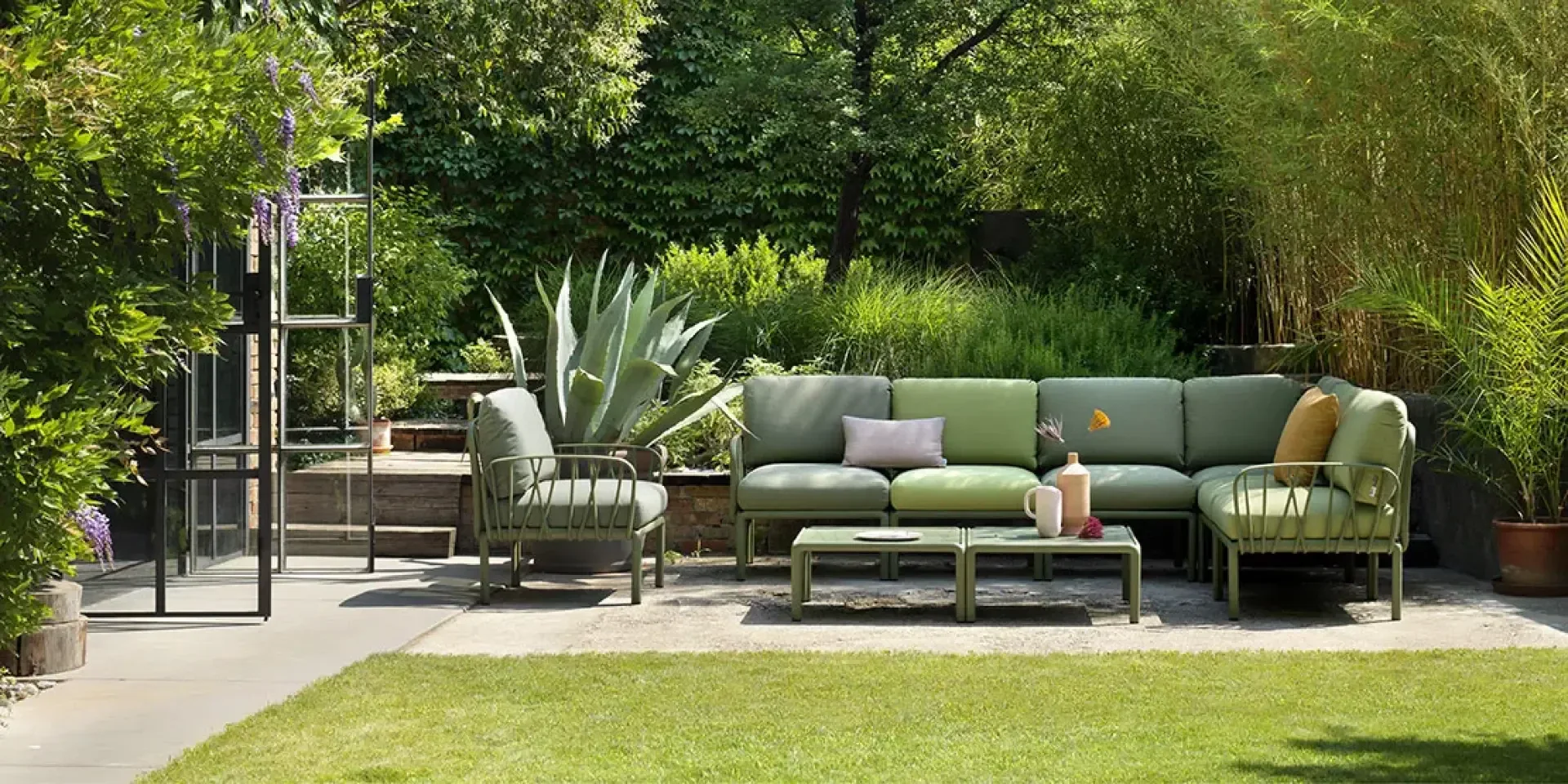 Outdoor patio with green cushioned sofa, armchairs, and a low table with vases. Surrounded by lush green plants and trees, the seating area sits on a patio next to a glass door, creating a relaxing garden atmosphere.