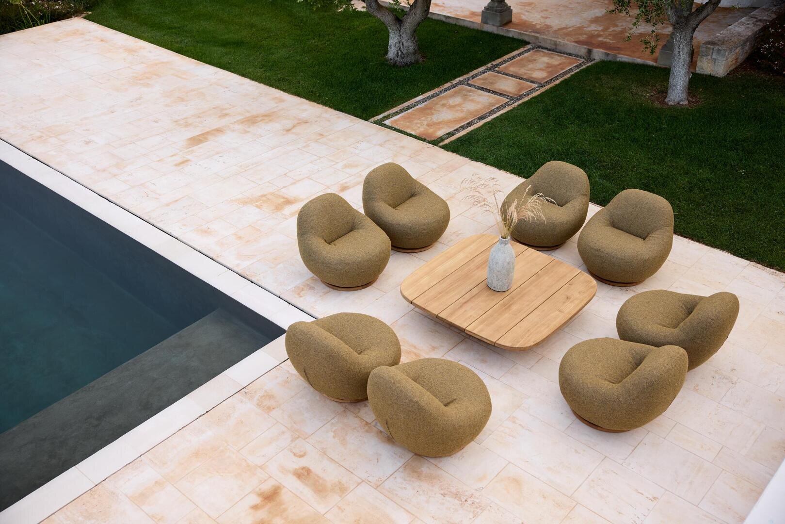 Eine Terrasse am Pool mit acht runden braunen Stühlen, die kreisförmig um einen quadratischen Holztisch mit einer Vase mit getrockneten Gräsern angeordnet sind und auf hellen Steinfliesen neben einer Grasfläche mit Bäumen stehen.