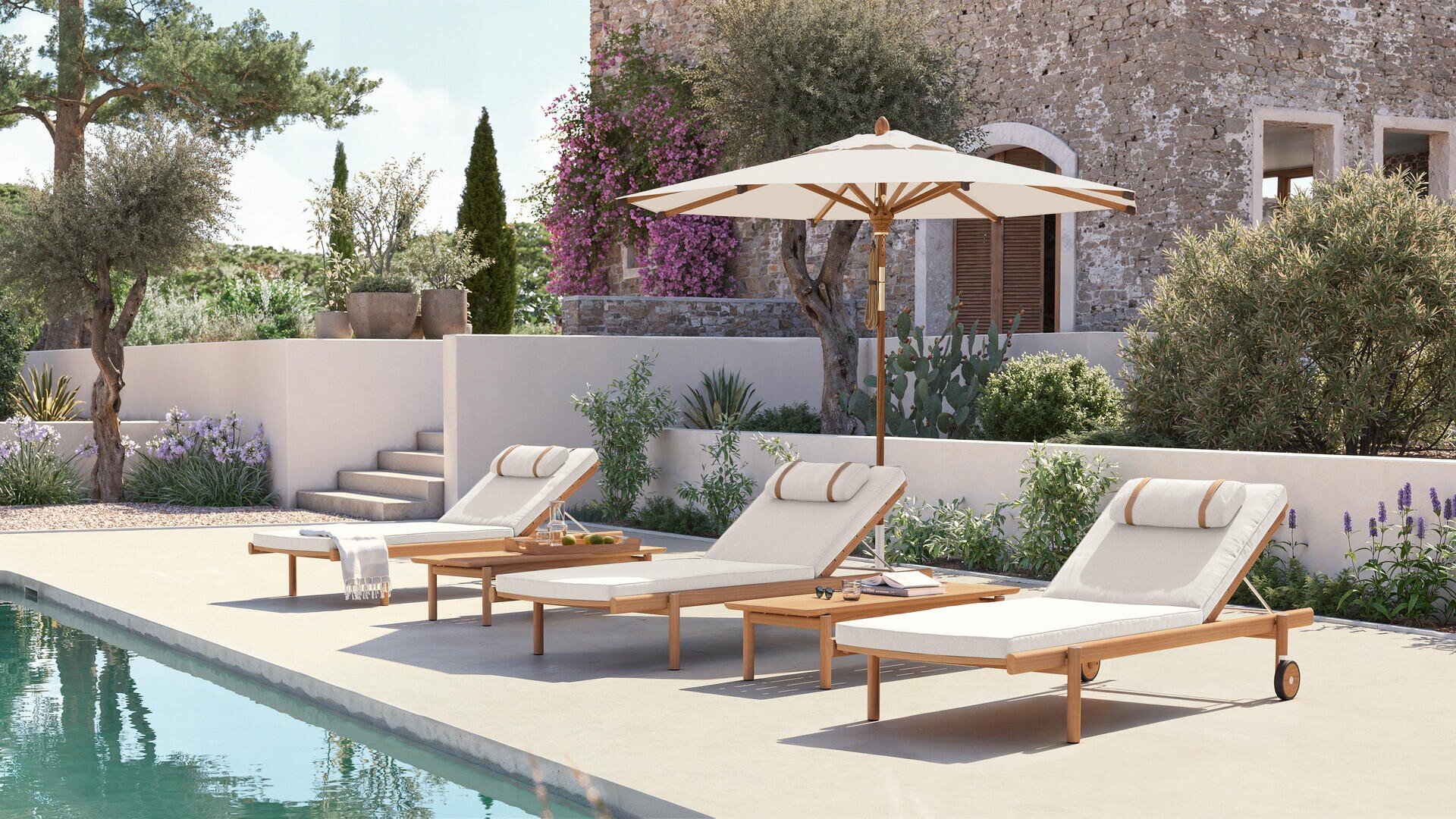 Drei gepolsterte Liegestühle mit gerollten Kopfstützen stehen am Pool, der von einem großen weißen Sonnenschirm beschattet wird. Im Hintergrund befinden sich Steinmauern, Treppen, Topfpflanzen und eine üppige Landschaftsgestaltung.