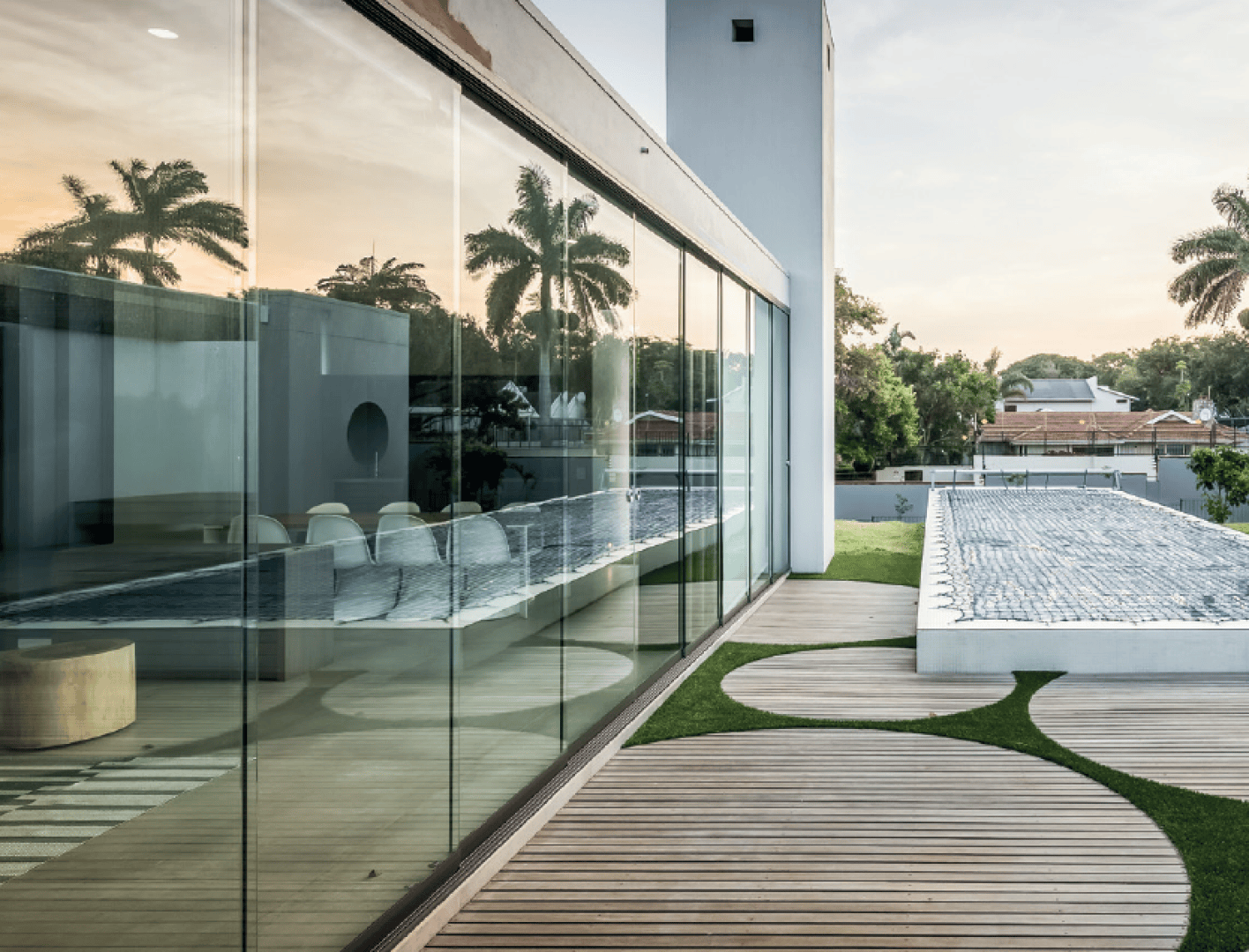 Modernes Haus mit großen Glasfenstern, in denen sich Palmen spiegeln, einer Holzterrasse mit kreisförmigem Grasmuster und einem rechteckigen Swimmingpool neben dem Gebäude bei Sonnenuntergang.