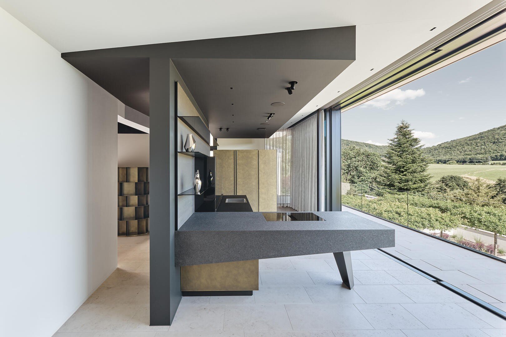A modern kitchen with minimalist gray cabinets, a smooth, floating worktop, open shelves with decorative items and large sliding glass doors overlooking a picturesque landscape of green hills and trees.