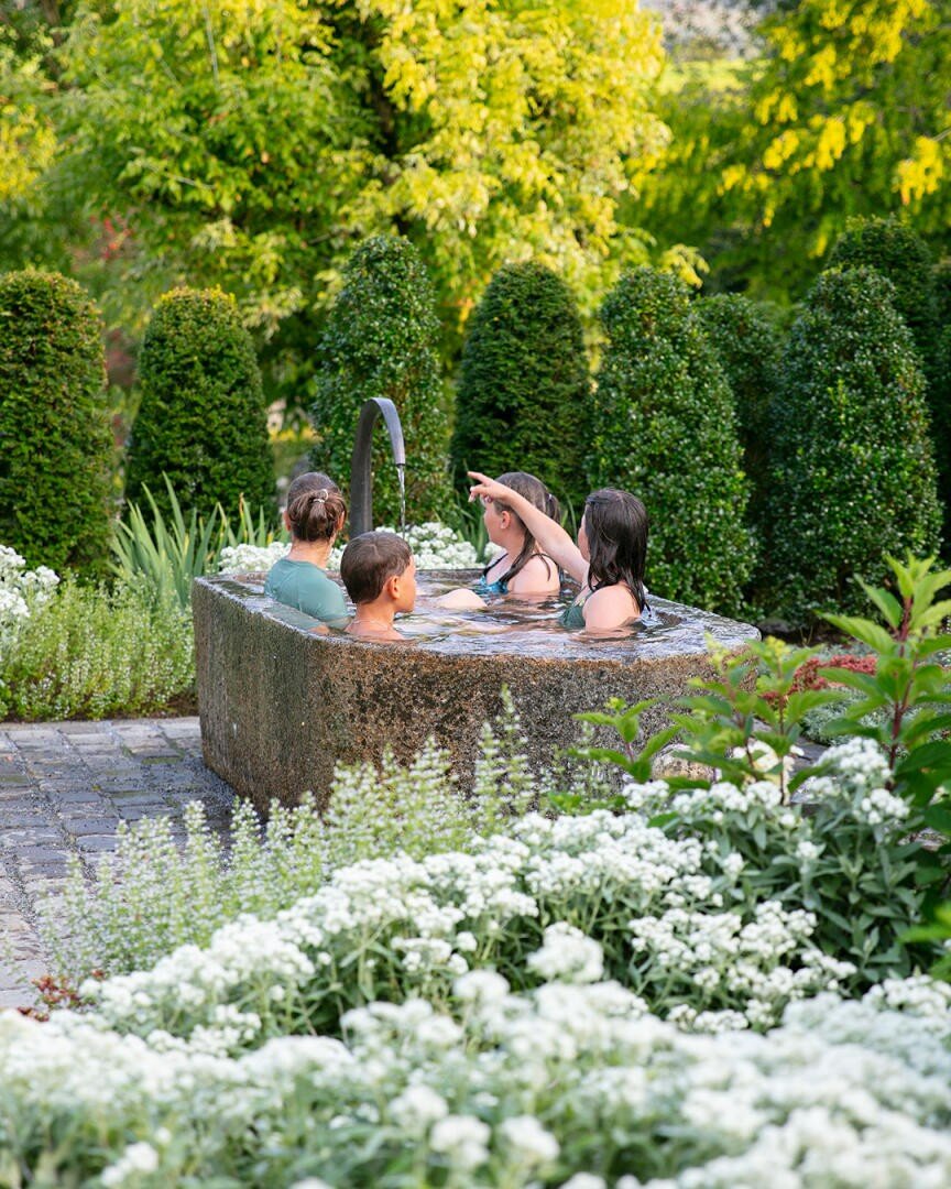Vier Personen entspannen sich an einem sonnigen Tag in einem kleinen Whirlpool aus Stein, umgeben von üppigem Grün, weißen Blumen und hohen Sträuchern in einer Gartenanlage.