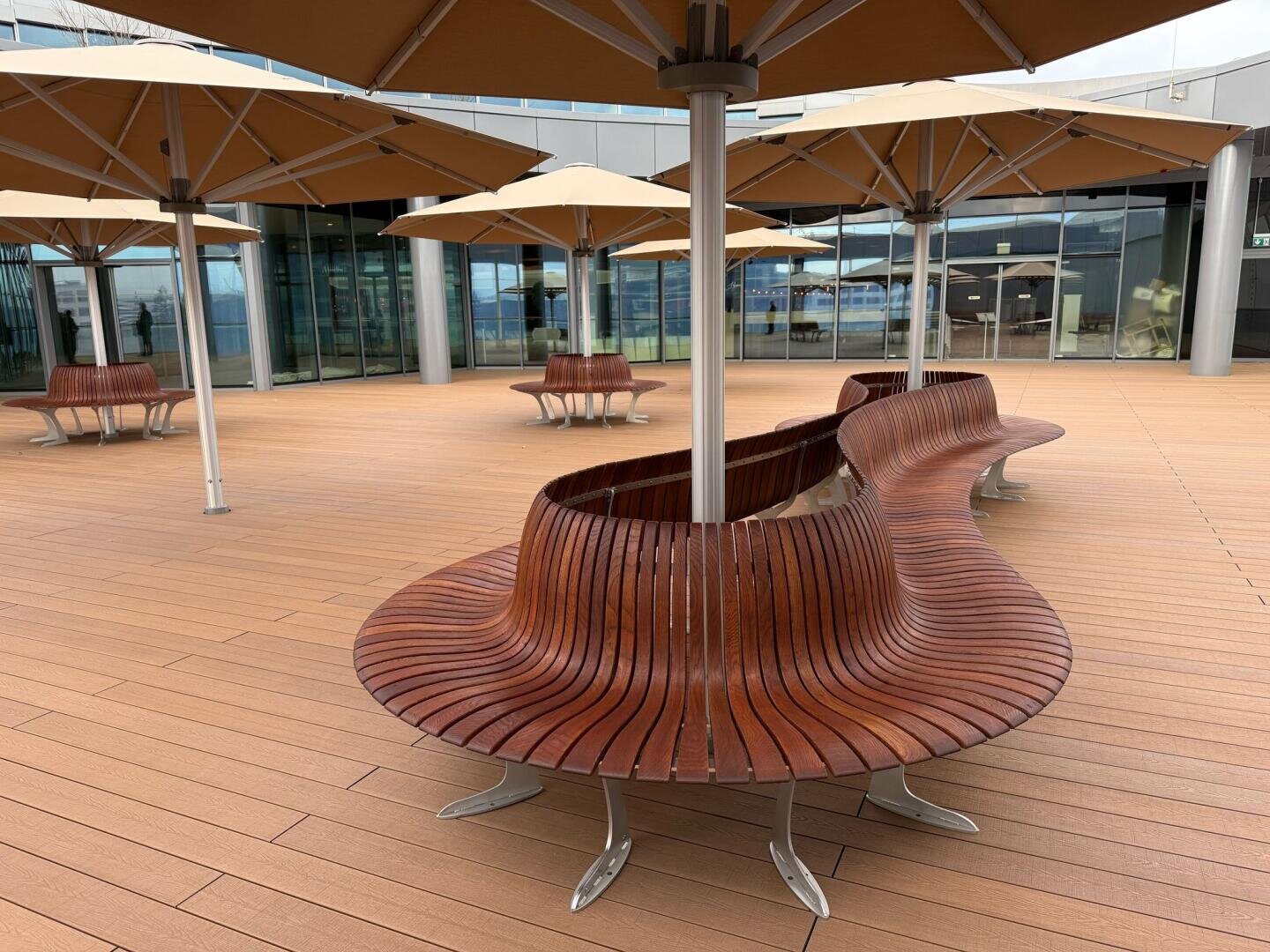 Curved wooden benches surround parasol poles on a spacious outdoor terrace with light brown flooring, large brown parasols and glass building entrances in the background.