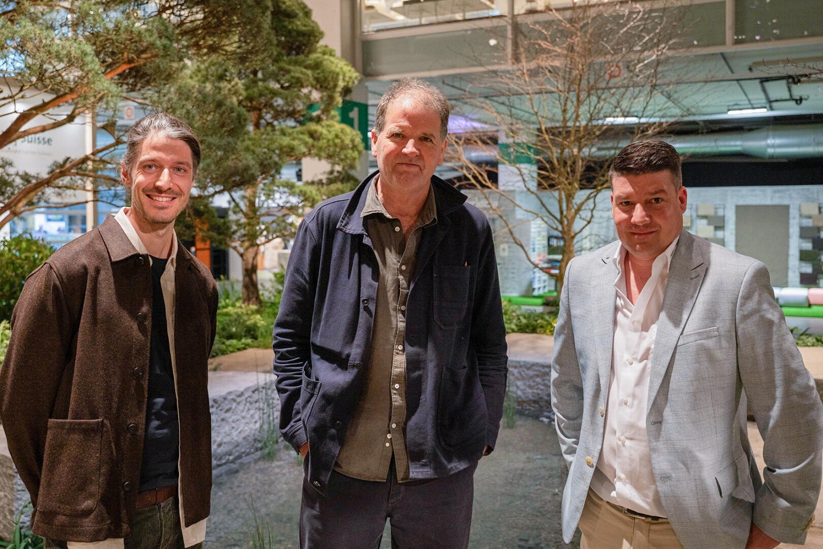 Drei Männer stehen zusammen in einem Innenraum vor einer Grünanlage mit Bäumen und Pflanzen. Alle tragen Jacken und lächeln leicht, im Hintergrund ist eine moderne Einrichtung zu sehen.