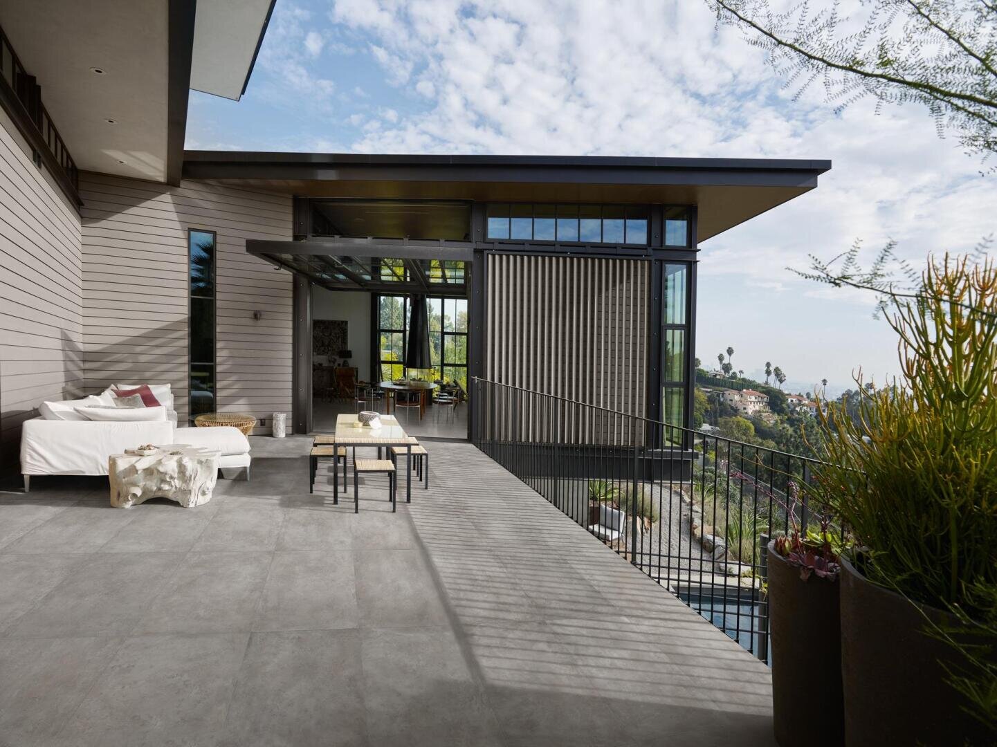 Modernes Haus mit großen Fenstern und geräumiger Terrasse mit Sitzgelegenheiten im Freien, Tischen und üppigen Pflanzen; Blick auf einen malerischen Hügel mit Bäumen unter einem teilweise bewölkten Himmel.