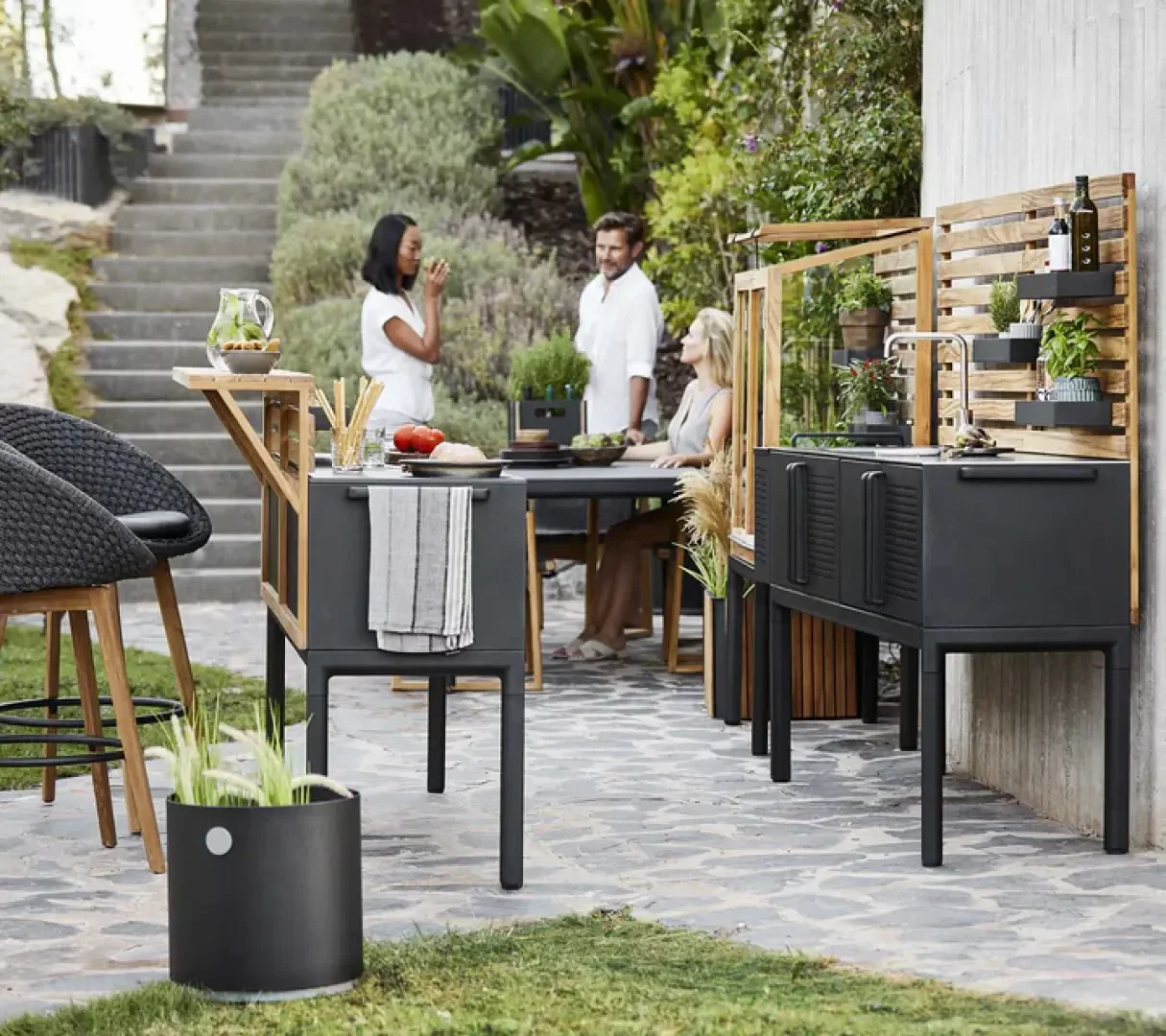 Drei Personen unterhalten sich in einer modernen Außenküche und einem Essbereich, umgeben von Grün und stilvollen schwarzen Möbeln auf einer Steinterrasse.