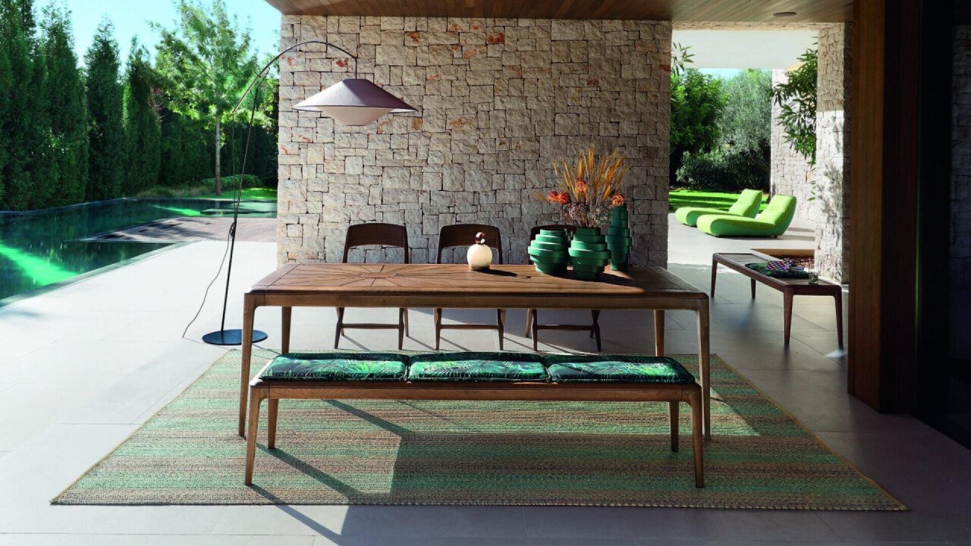 A modern outdoor dining area features a wooden table with green vases, chairs, a cushioned bench, floor lamp, and a woven rug beside a stone wall. A pool and green lounge chairs are visible in the background.