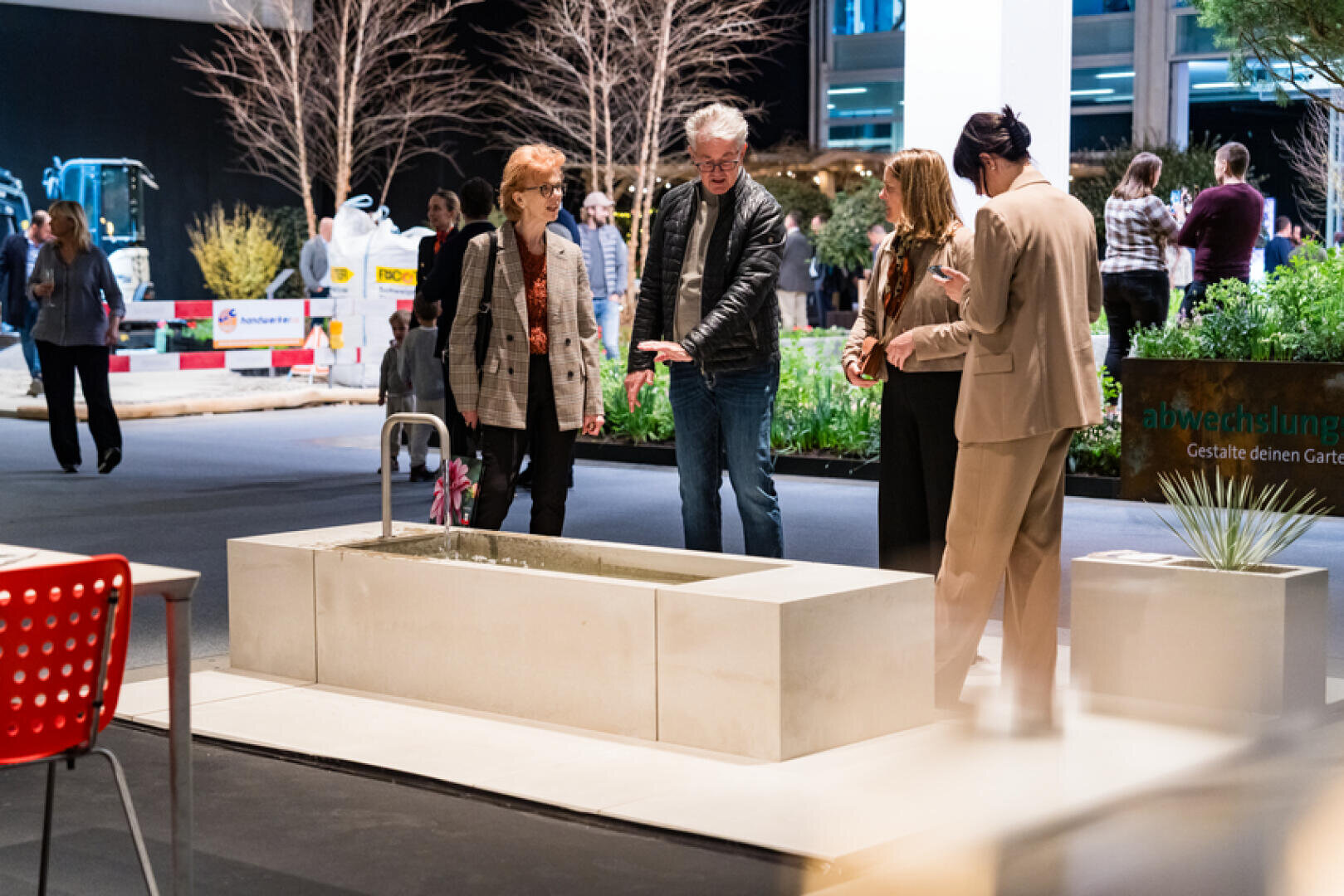 Vier Erwachsene stehen in einer Indoor-Ausstellung um ein modernes Wasserspiel im Freien herum, diskutieren und gestikulieren. Im Hintergrund sind andere Menschen und Gartenausstellungen zu sehen.