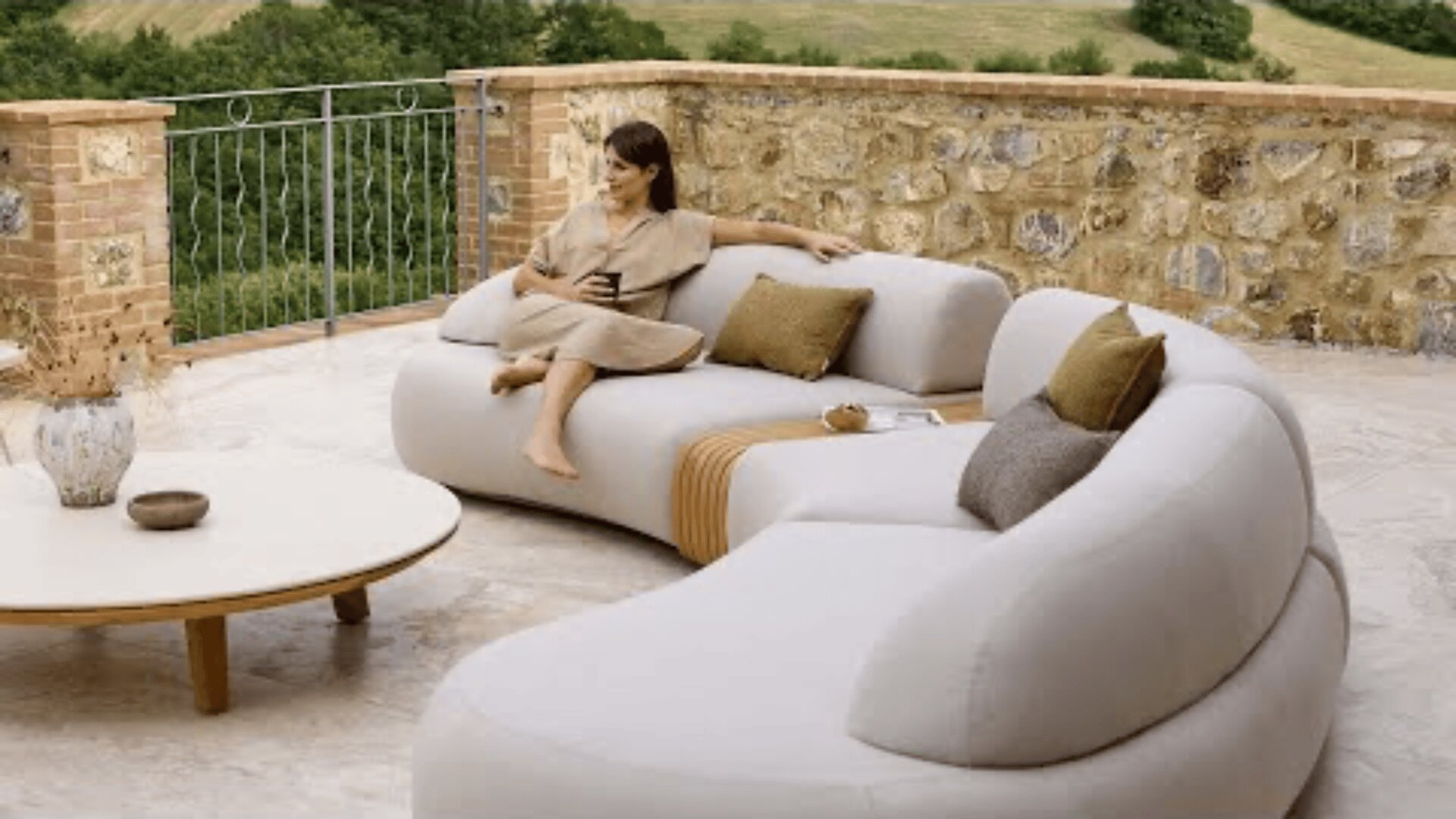 A woman in a beige dress sits on a modern curved outdoor sofa with brown cushions on a stone patio, holding a drink. A round coffee table and a scenic countryside view are in the background.