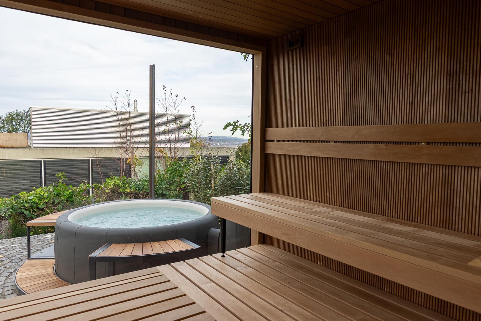 The interior of the wooden sauna with its tiered benches overlooks a garden with a round whirlpool surrounded by greenery and a terrace that can be seen through a large open window.