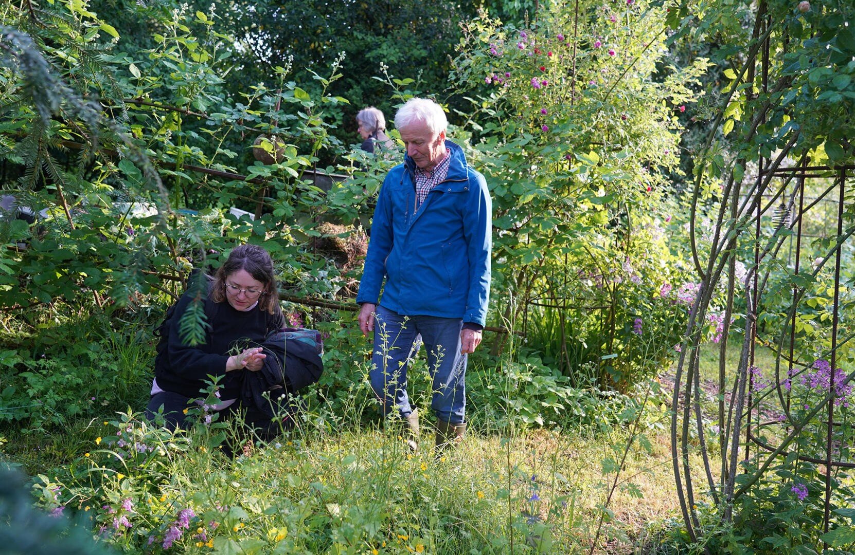 Zwei Personen befinden sich in einem üppigen, grünen Garten. Eine Person hockt und untersucht die Pflanzen, während die andere daneben steht. Eine weitere Person ist im Hintergrund inmitten des Grüns und der Blumen zu sehen.