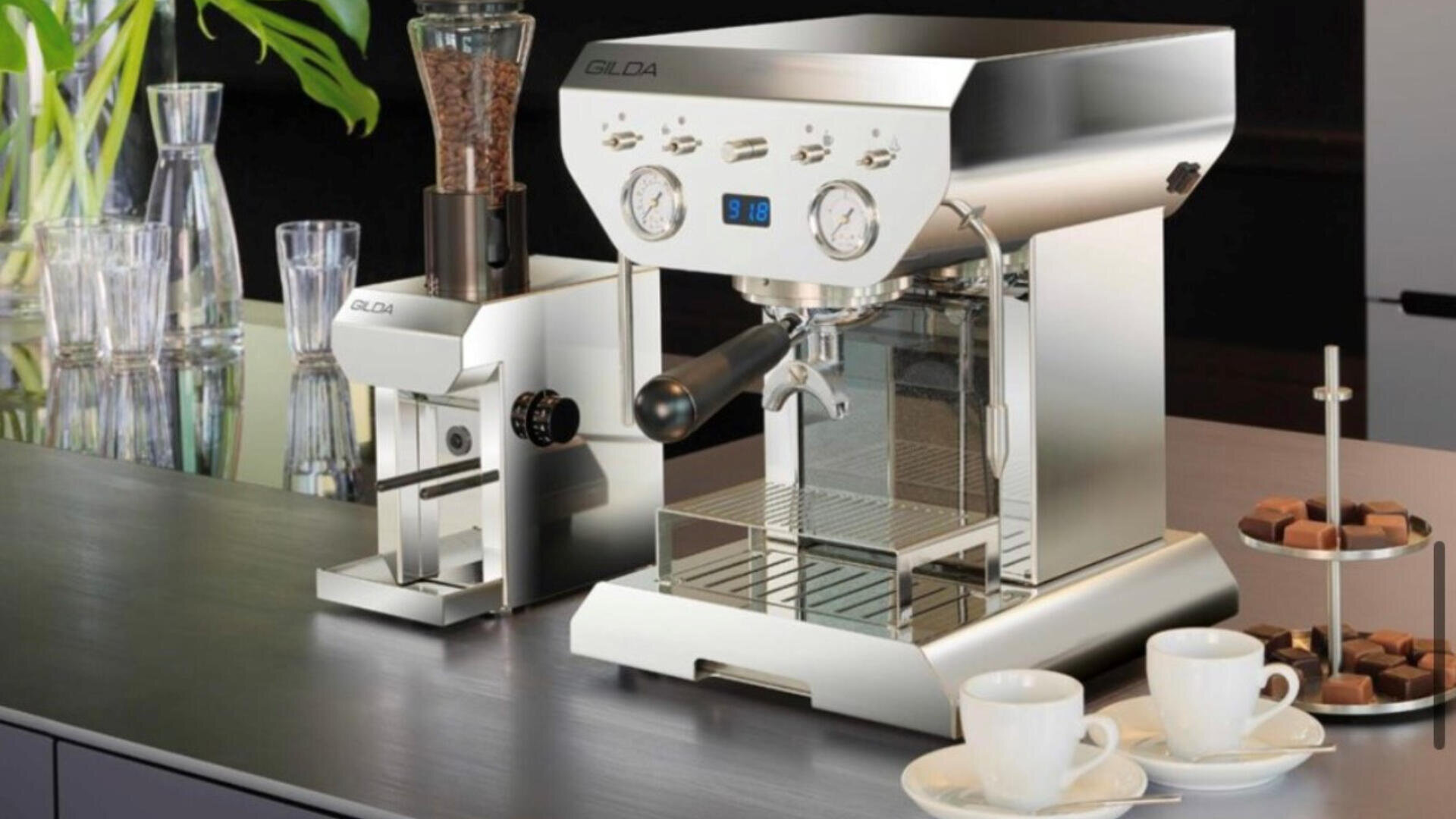 Eine glänzende Espressomaschine und eine Kaffeemühle stehen auf einem modernen Küchentisch, umgeben von Tassen, Gläsern, einem Teller mit Pralinen und Grünpflanzen im Hintergrund.
