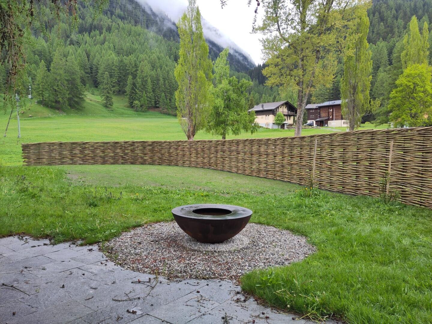A round metal fireplace lies on gravel in a grassy courtyard bordered by a woven wooden fence. Behind the fence are green fields, trees and buildings, with wooded mountains in the background.