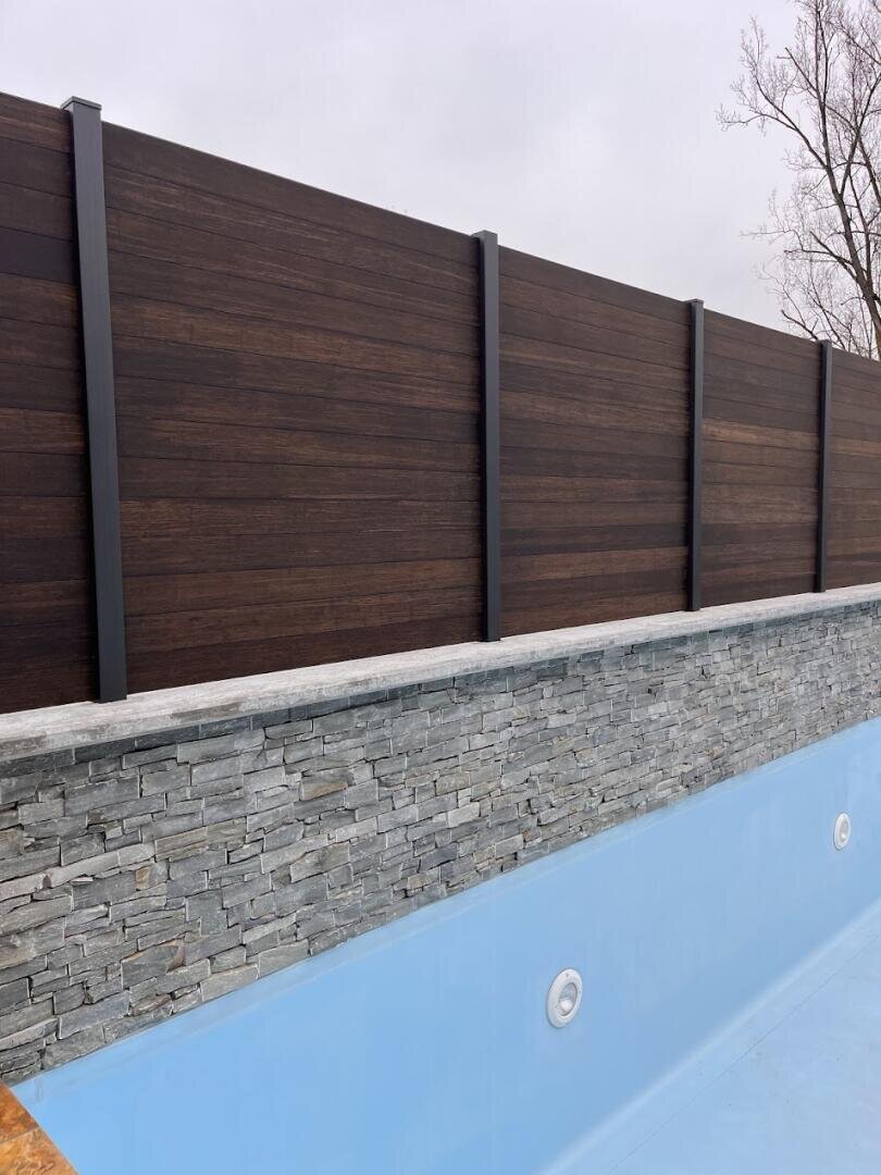 Leerer Swimmingpool mit hellblauem Innenraum, steingefliestem Rand und einem hohen, dunklen Holzzaun im Hintergrund. Hinter dem Zaun sind ein bedeckter Himmel und kahle Baumzweige zu sehen.