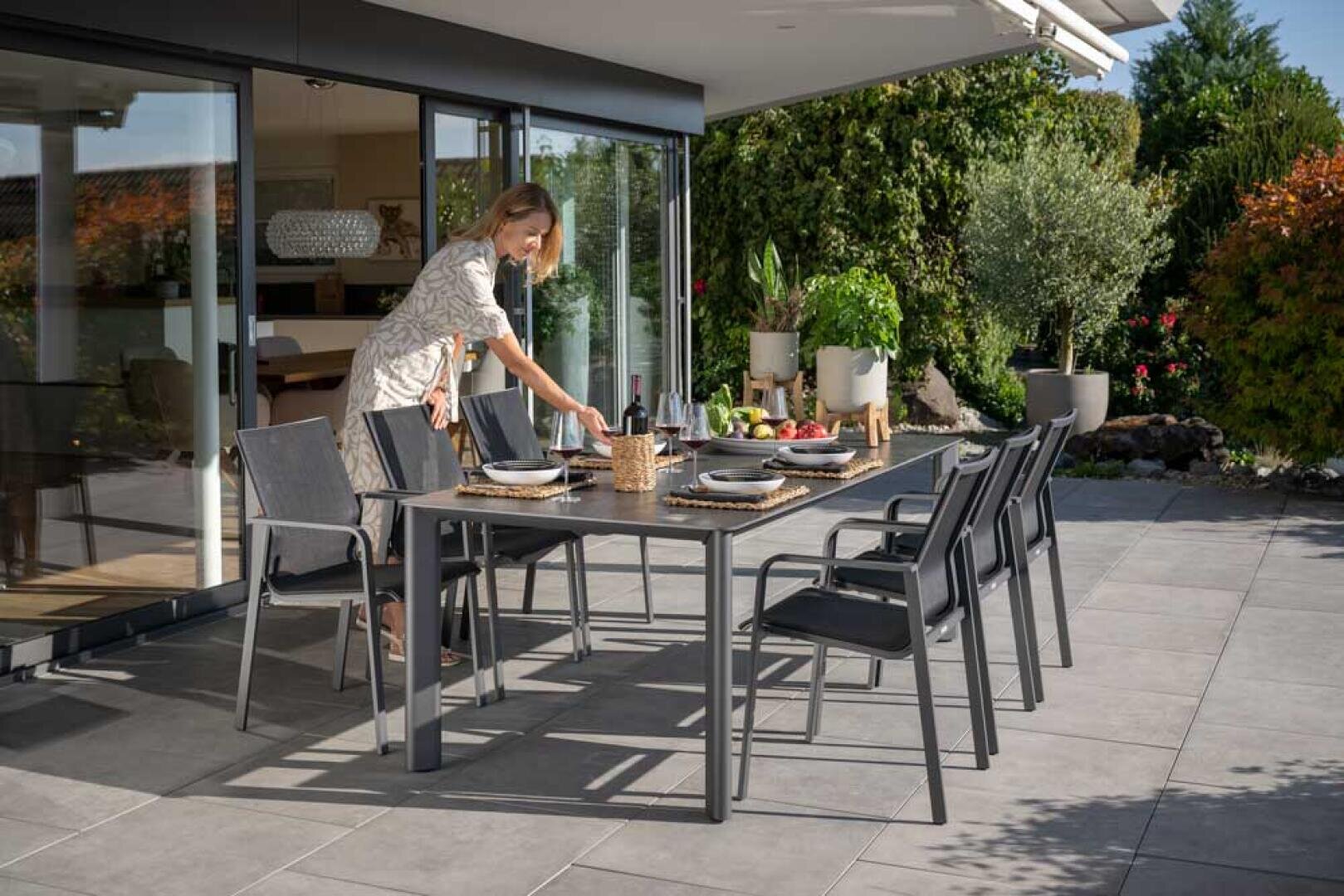 Eine Frau arrangiert Teller auf einem modernen Esstisch im Freien, der für eine Mahlzeit auf einer Terrasse gedeckt ist, umgeben von viel Grün und einem modernen Haus mit großen Glastüren.