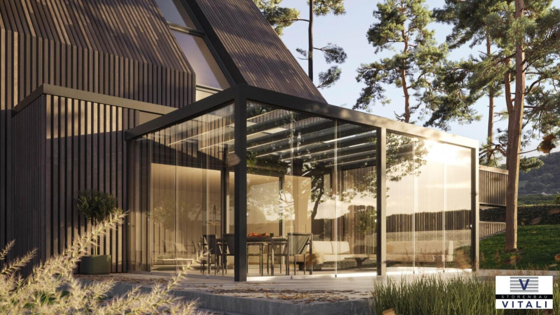 Modern home with dark wood exterior and large glass-walled patio enclosure. Inside the enclosure is a dining table and chairs. Surrounded by trees and greenery, with sunlight reflecting off the glass. Vitali logo in bottom right corner.