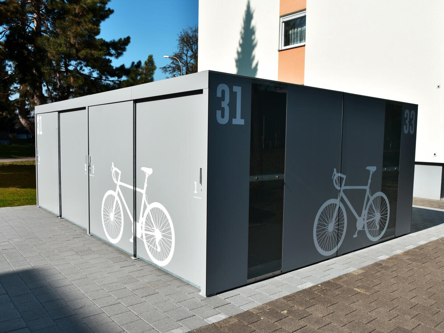 Ein moderner grauer Fahrradschuppen mit großen weißen Fahrradgrafiken und den Nummern 31 und 33 auf den Türen, der neben einem weißen Gebäude auf einer gepflasterten Fläche steht.