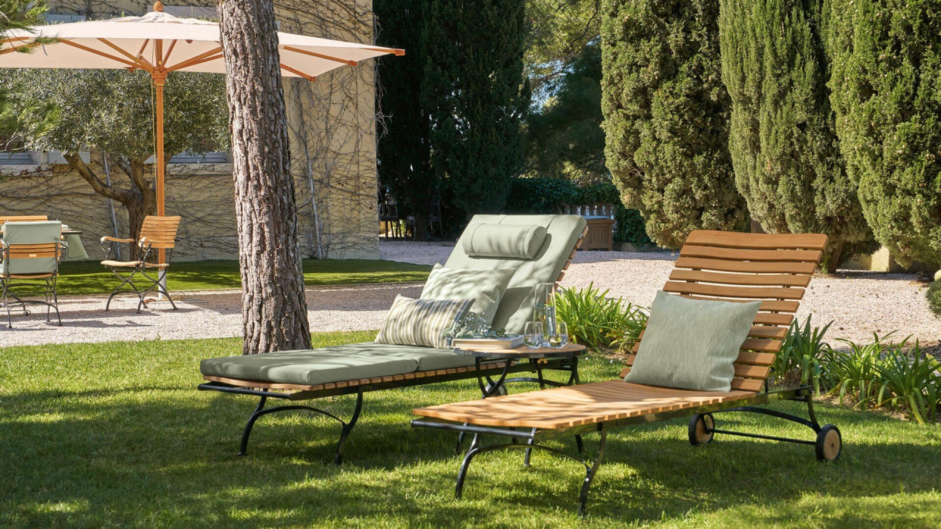 Zwei hölzerne Liegestühle mit Kissen sitzen auf einer Wiese in einem sonnigen Garten, mit Bäumen, einem Sonnenschirm und einem Tisch mit Stühlen im Hintergrund.
