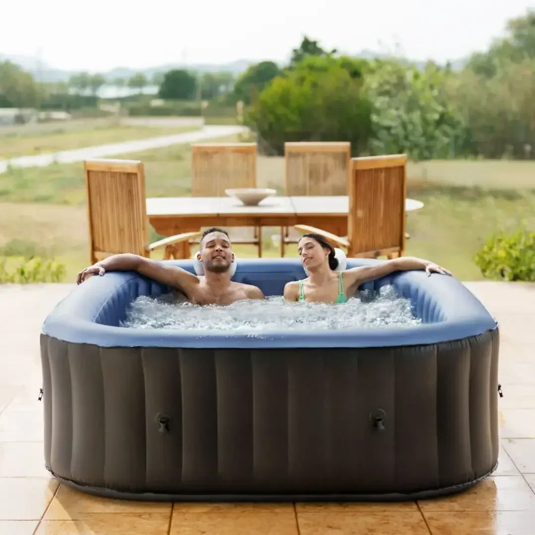 A man and a woman relax in an inflatable hot tub on a patio, smiling with their heads tilted back, surrounded by bubbling water. Outdoor dining furniture and trees are visible in the background.
