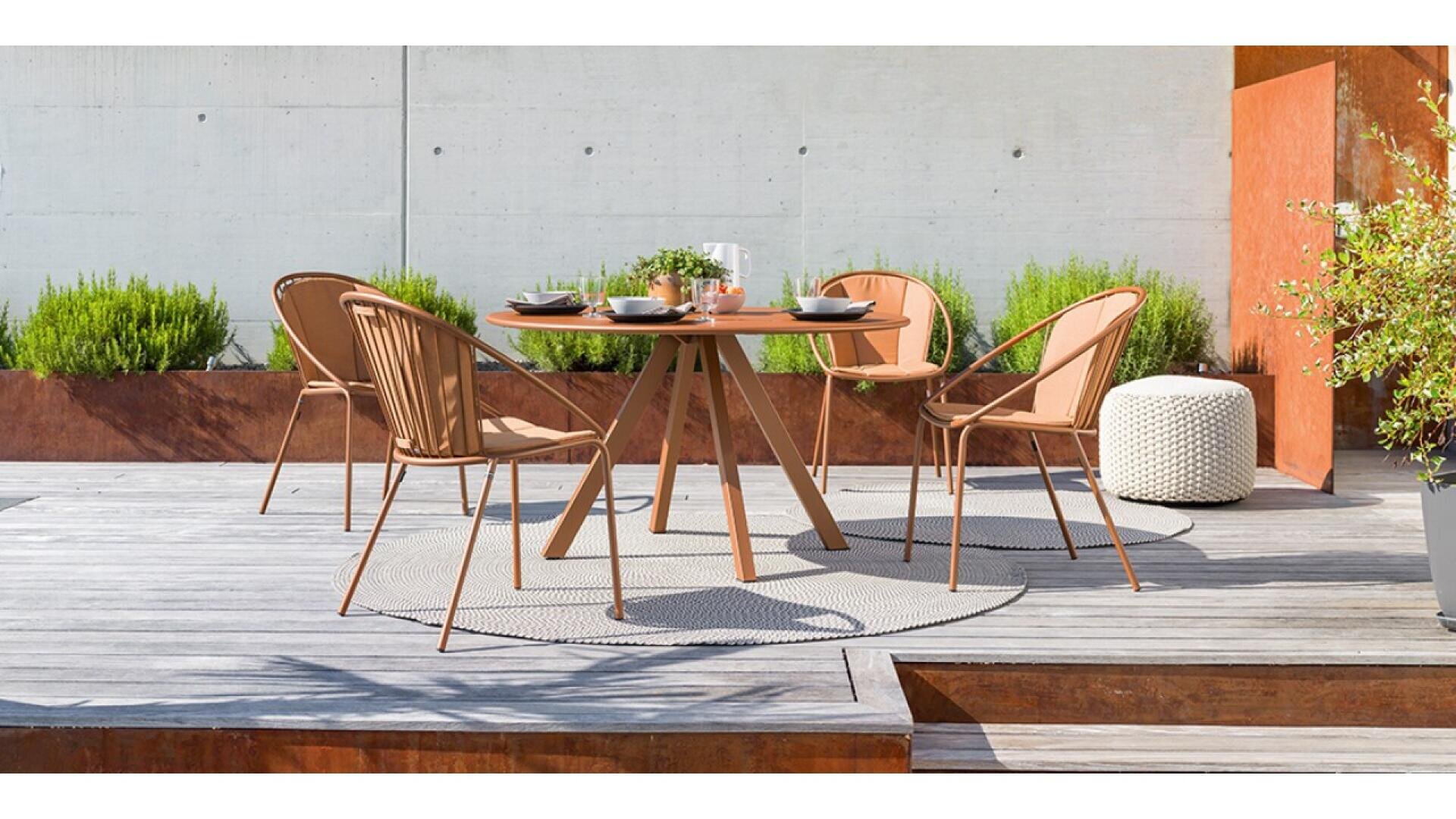 A modern outdoor dining set with a round wooden table, four tan chairs, tableware, and plants in the background, situated on a wooden deck with a light circular rug.