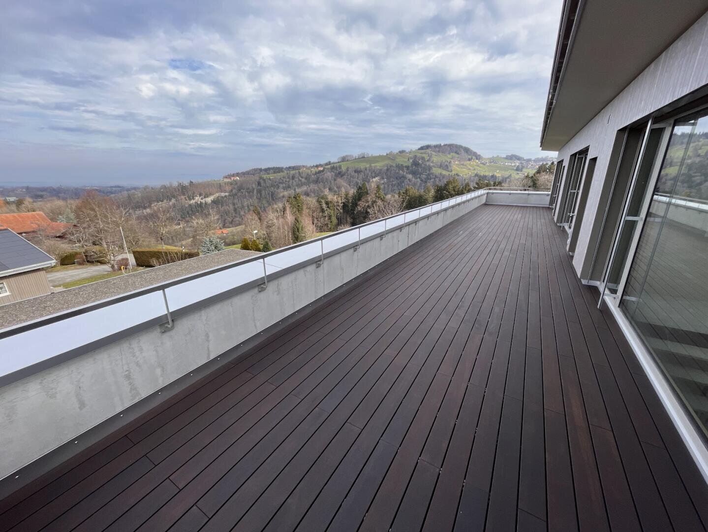 Ein geräumiger Balkon mit dunklem Holzfußboden und einem Metallgeländer bietet einen Blick auf eine malerische Landschaft mit Hügeln, Bäumen und Häusern unter einem teilweise bewölkten Himmel. Große Fenster säumen die rechte Seite des modernen Gebäudes.