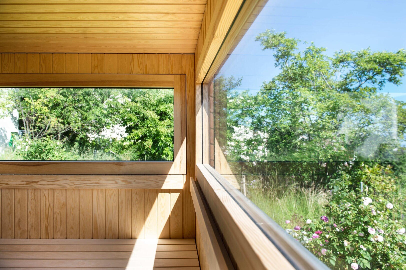 Eine helle, hölzerne Saunakabine mit großen Fenstern bietet einen Blick auf einen üppigen, grünen Garten mit Bäumen und blühenden Sträuchern unter einem klaren blauen Himmel. Das Sonnenlicht strömt in den Raum und schafft eine warme und einladende Atmosphäre.
