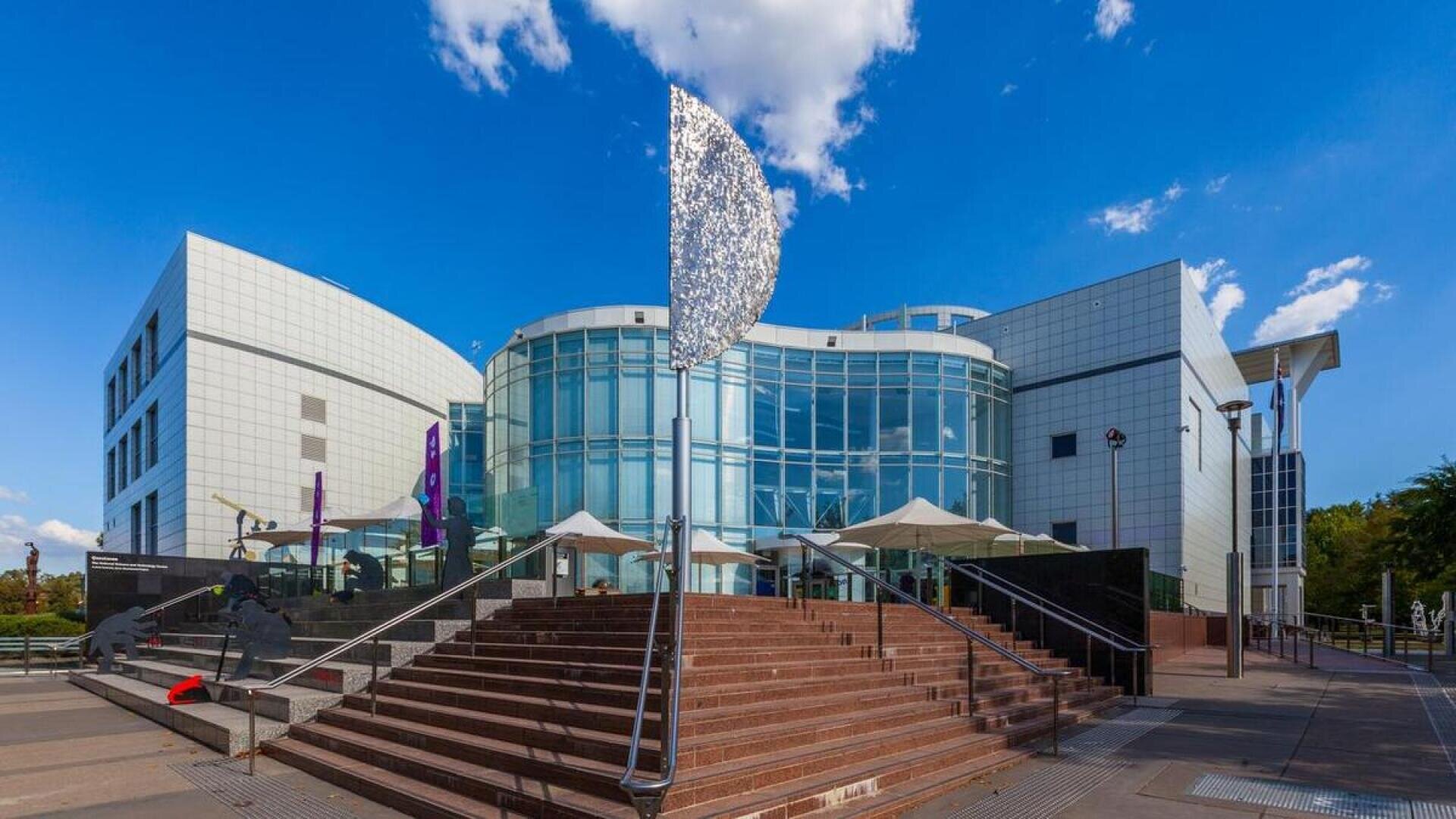 Modern building with a glass façade and white panels, wide steps leading up to the entrance, a metal sculpture in front, clear blue sky with a few clouds overhead.