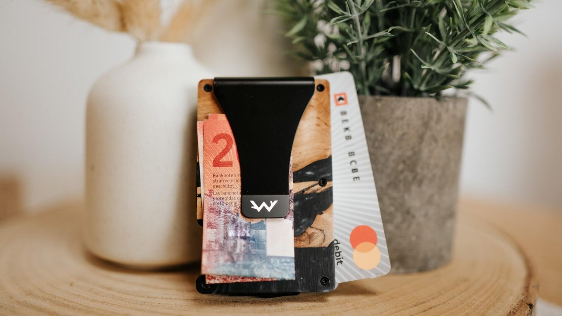 Ein minimalistisches Portemonnaie enthält eine gefaltete rote Banknote und zwei Karten, darunter eine Mastercard-Debitkarte, die auf einer Holzfläche neben dekorativen Pflanzen in Vasen stehen.